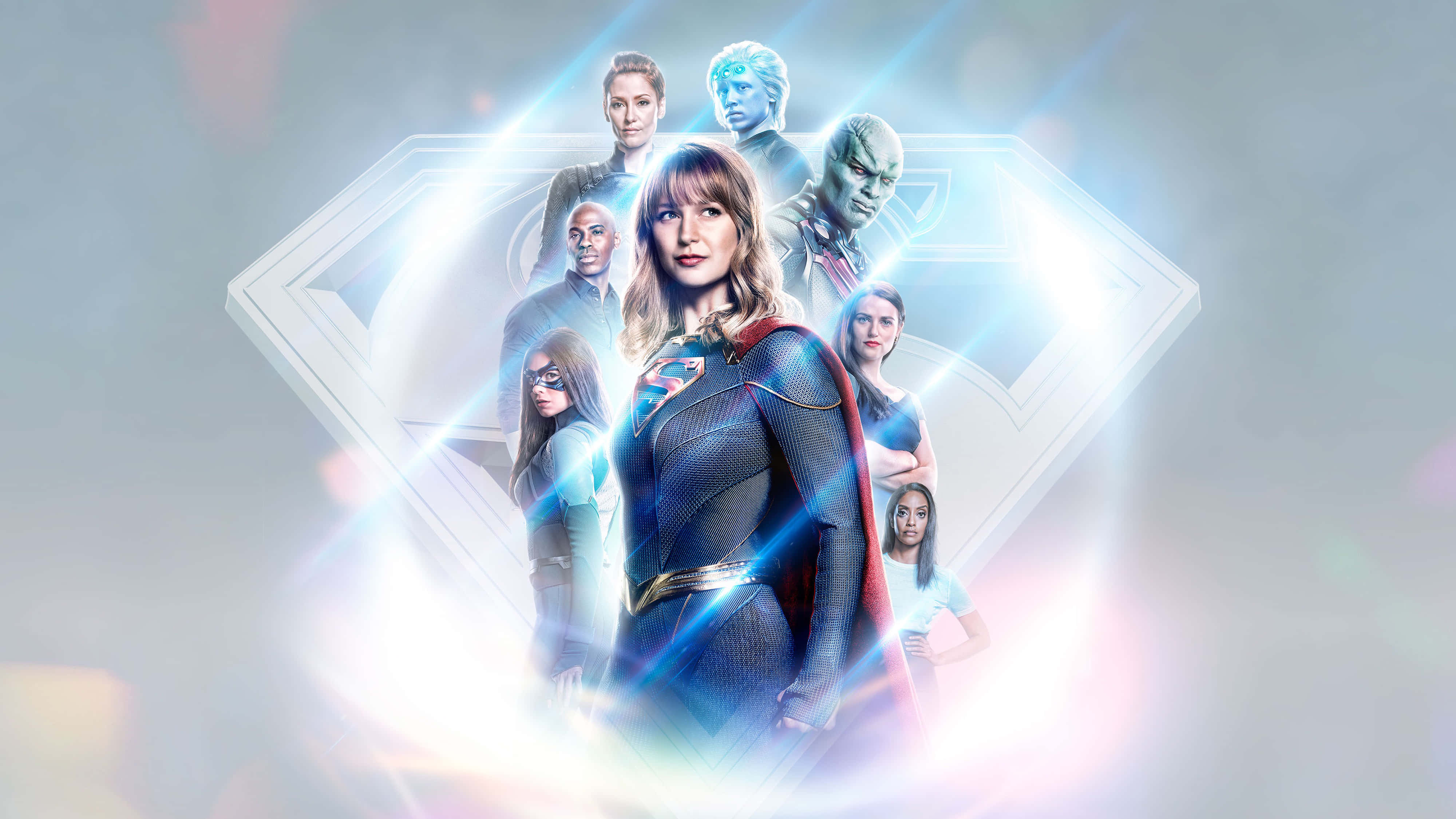 Chyler Leigh 桌布 壁紙 — 一組光芒四射的超級英雄站在發光的超人標誌內，沐浴在明亮的藍色和白色光線中。