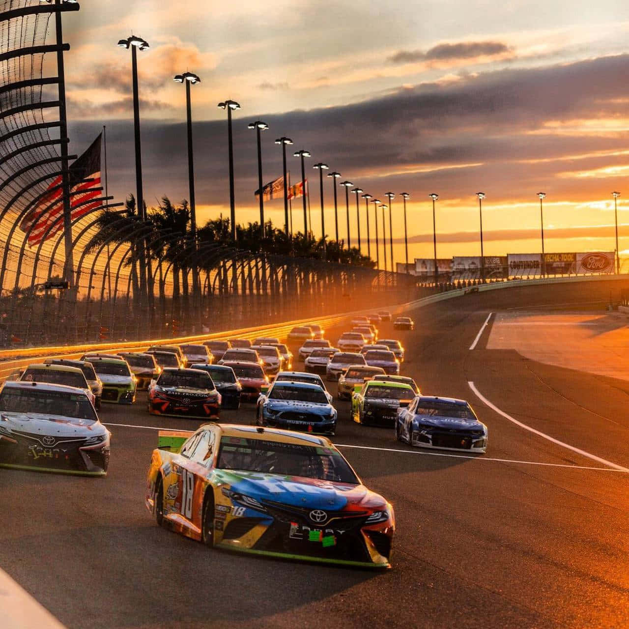 nascar 桌布 壁紙 — 一組色彩繽紛的NASCAR賽車在日落時分繞著賽道飛馳，背景有棕櫚樹和美國國旗。