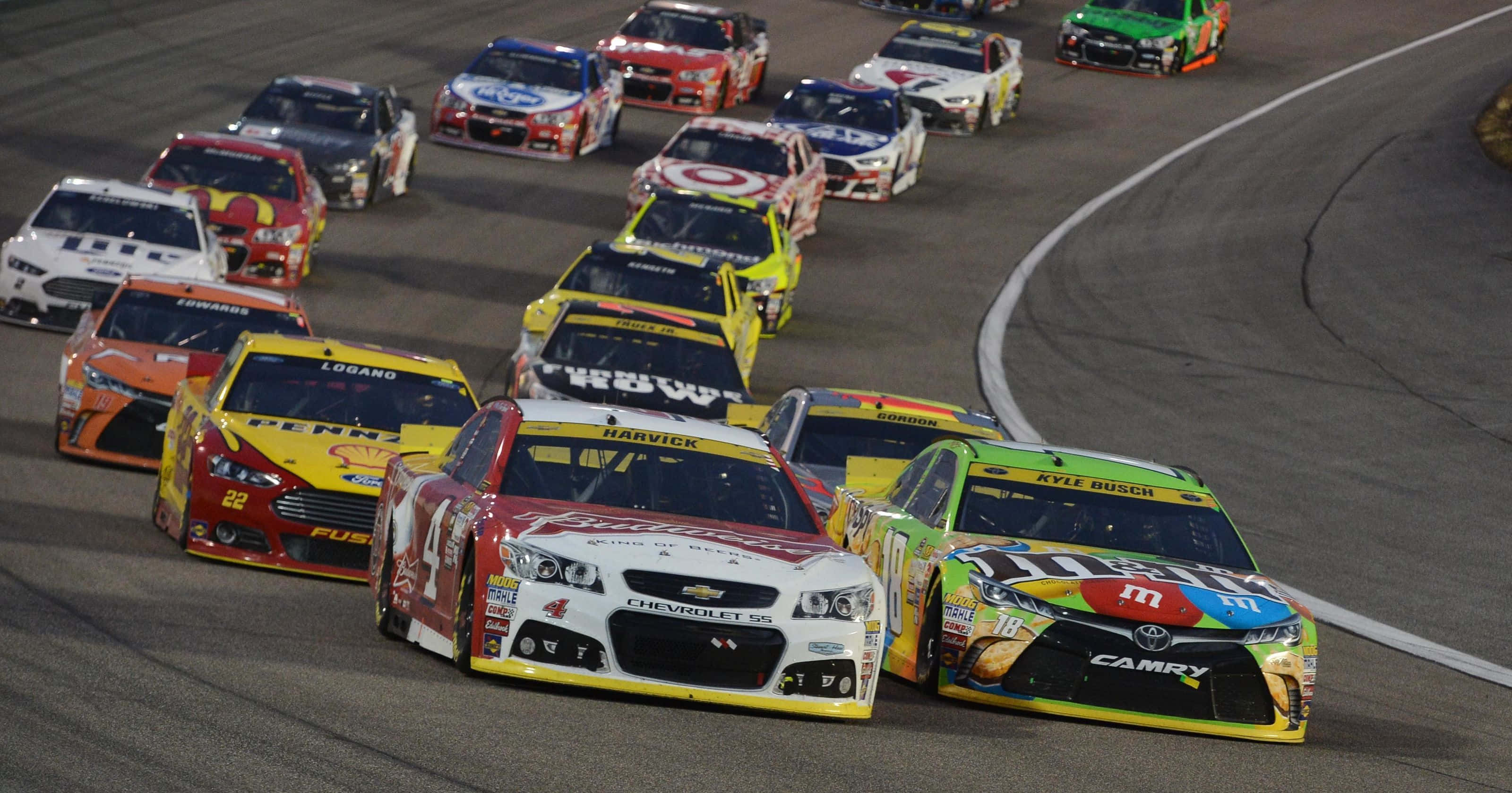 nascar 桌布 壁紙 — 一組色彩鮮豔的NASCAR賽車在夜間瀝青彎道上，在體育場燈光下高速飛馳。