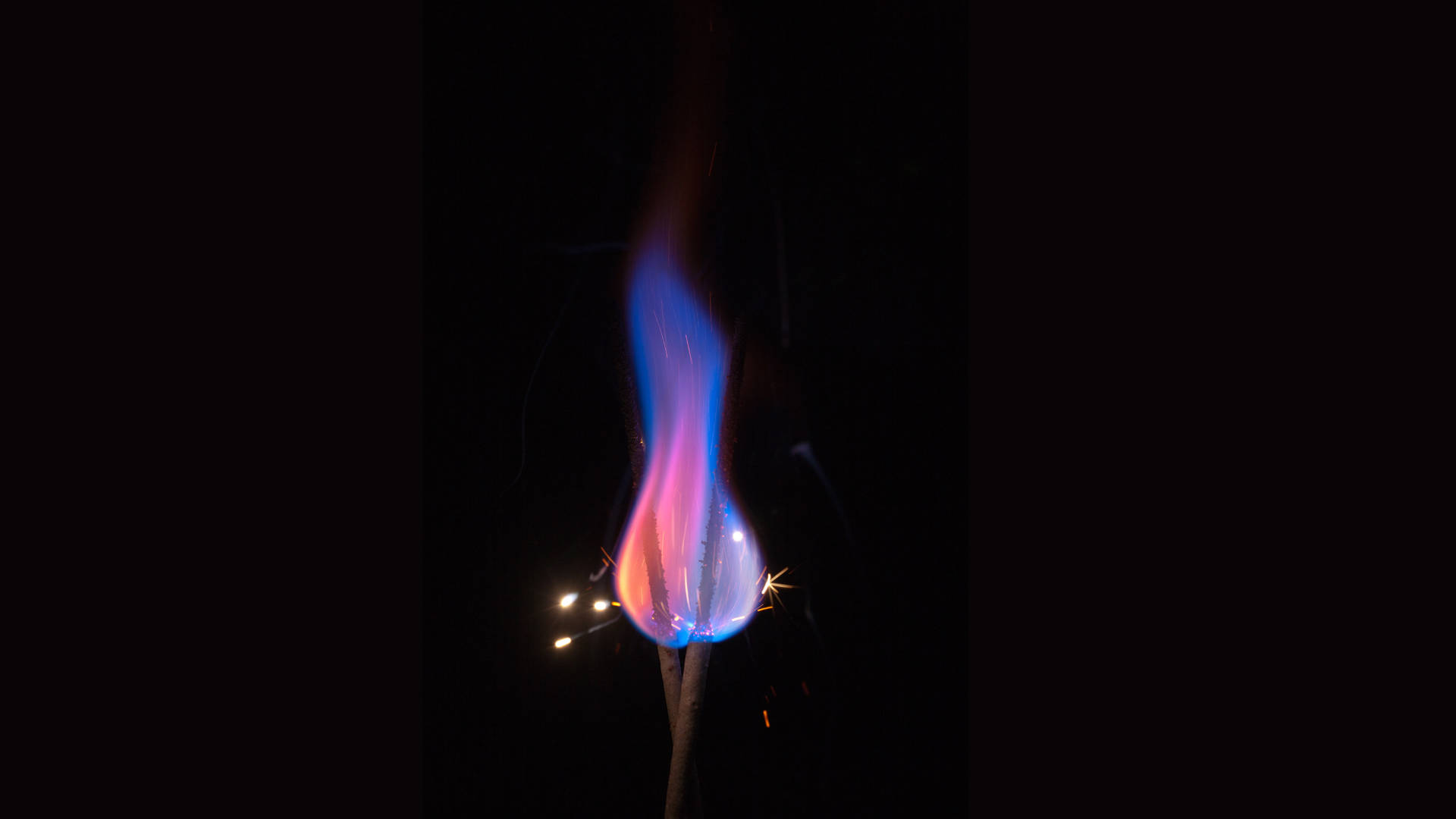 Flames 桌布 壁紙 — 一縷充滿活力的藍色和粉紅色火焰在漆黑的背景上向上舞動，下方有發光的火花拖曳。