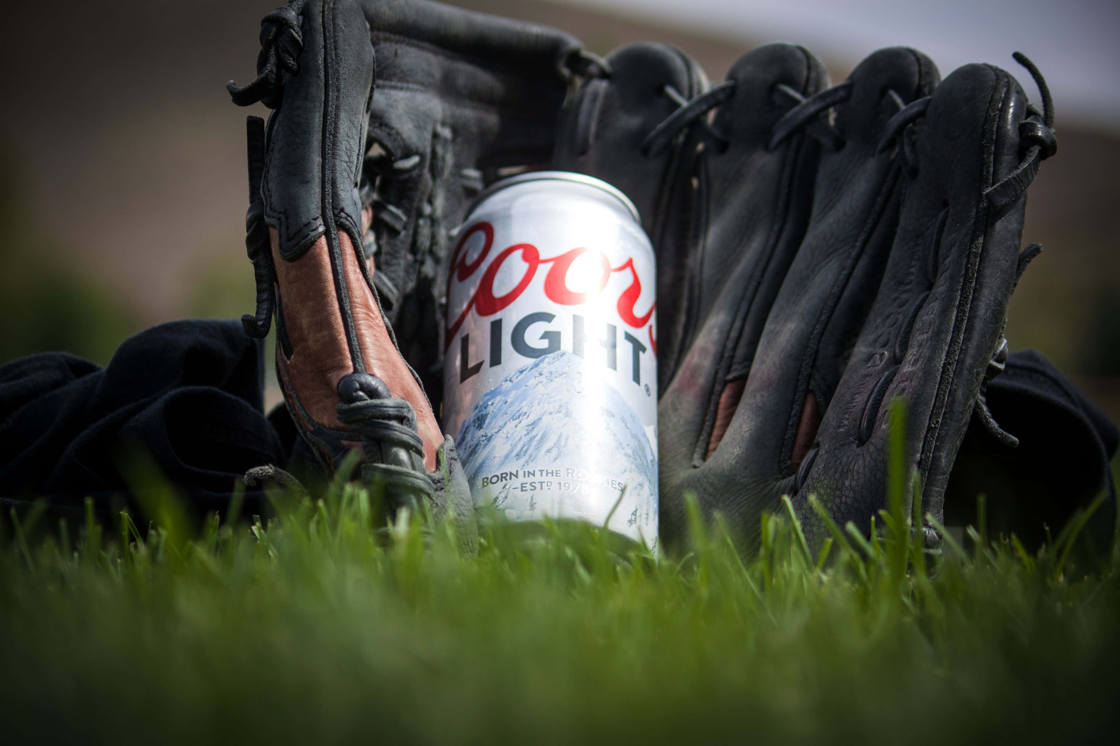 Beer Can 桌布 壁紙 — 一罐Coors Light啤酒放在磨損的棒球手套和釘鞋之間，在鮮綠的草地上。