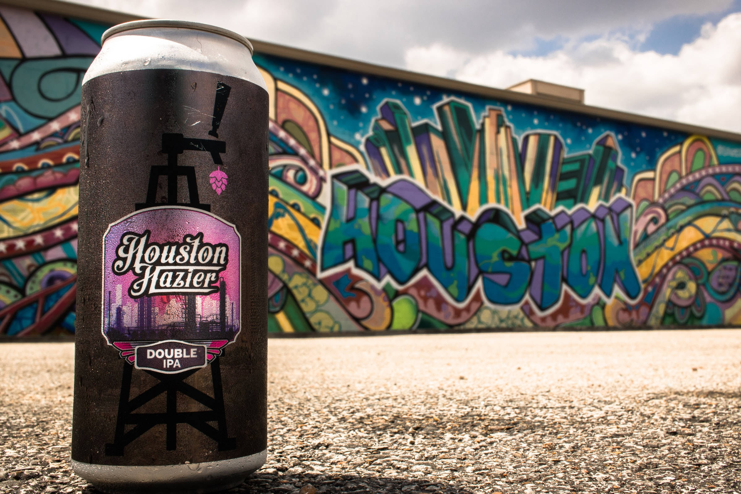 Houston 桌布 壁紙 — 一罐冰涼的Houston Hazier Double IPA啤酒放在人行道上，前面是充滿活力的塗鴉壁畫，在陰天天空下拼出Houston。