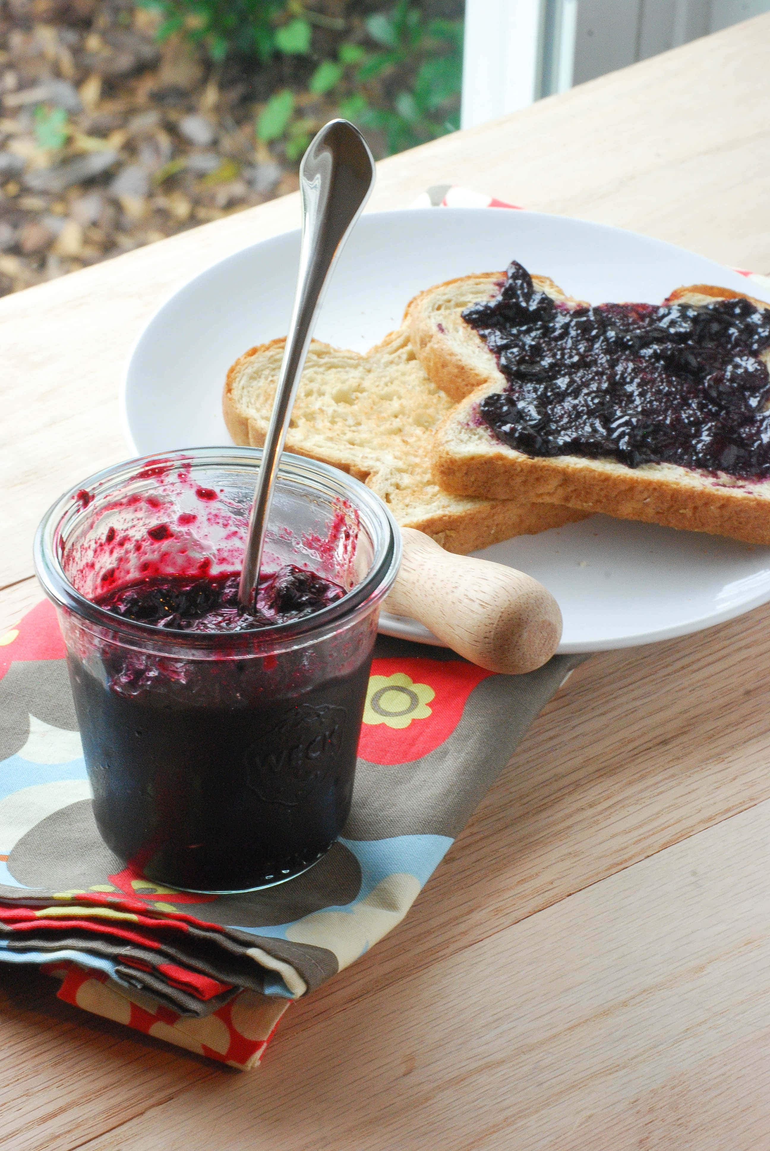 Blueberry Jam 桌布 壁紙 — 一罐深色莓果醬配湯匙，放在木桌上色彩繽紛的餐巾上的盤子旁，旁邊有吐司。