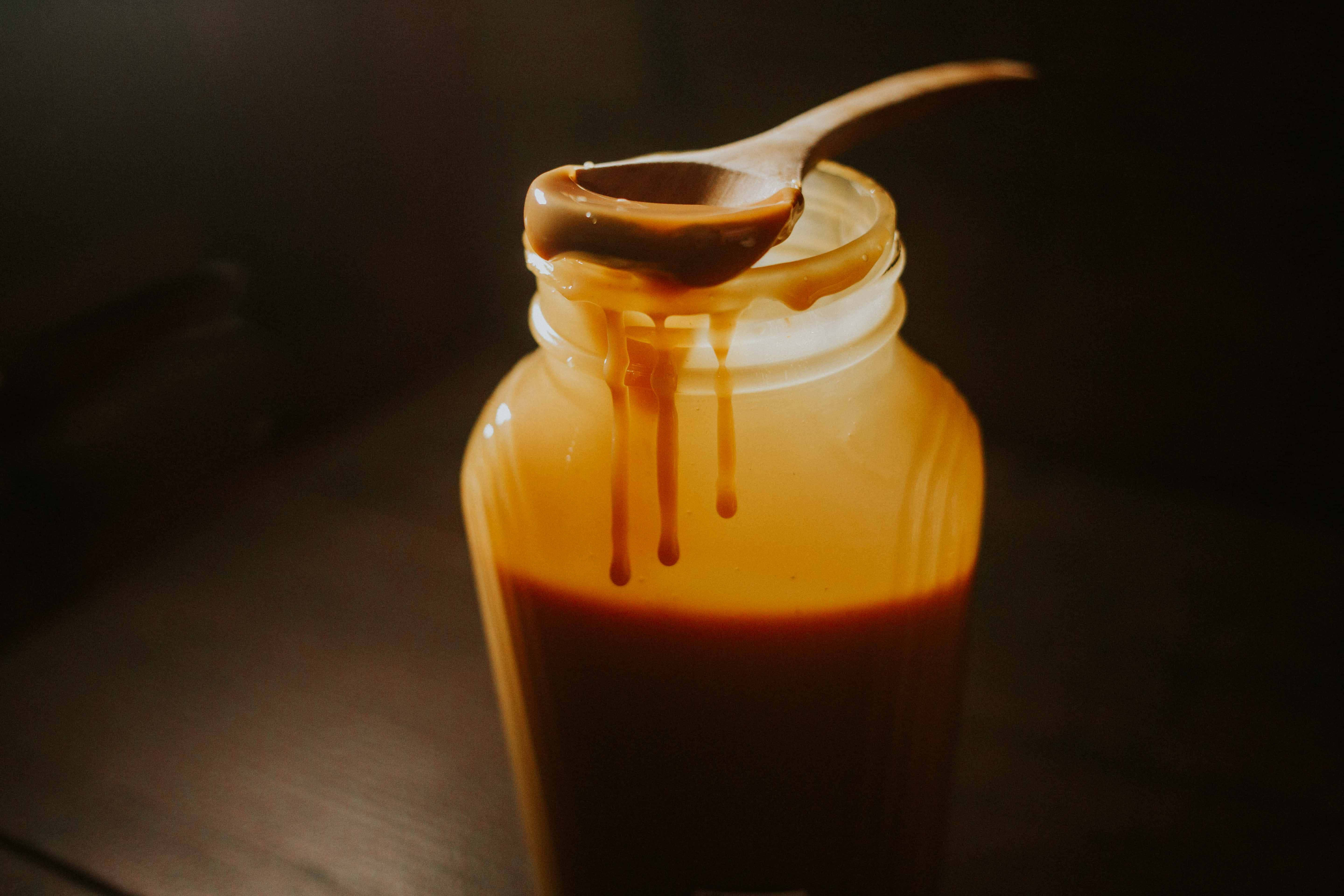 Caramel 桌布 壁紙 — 一罐濃郁的焦糖醬，搭配木勺在深色背景下滴落金色糖漿。