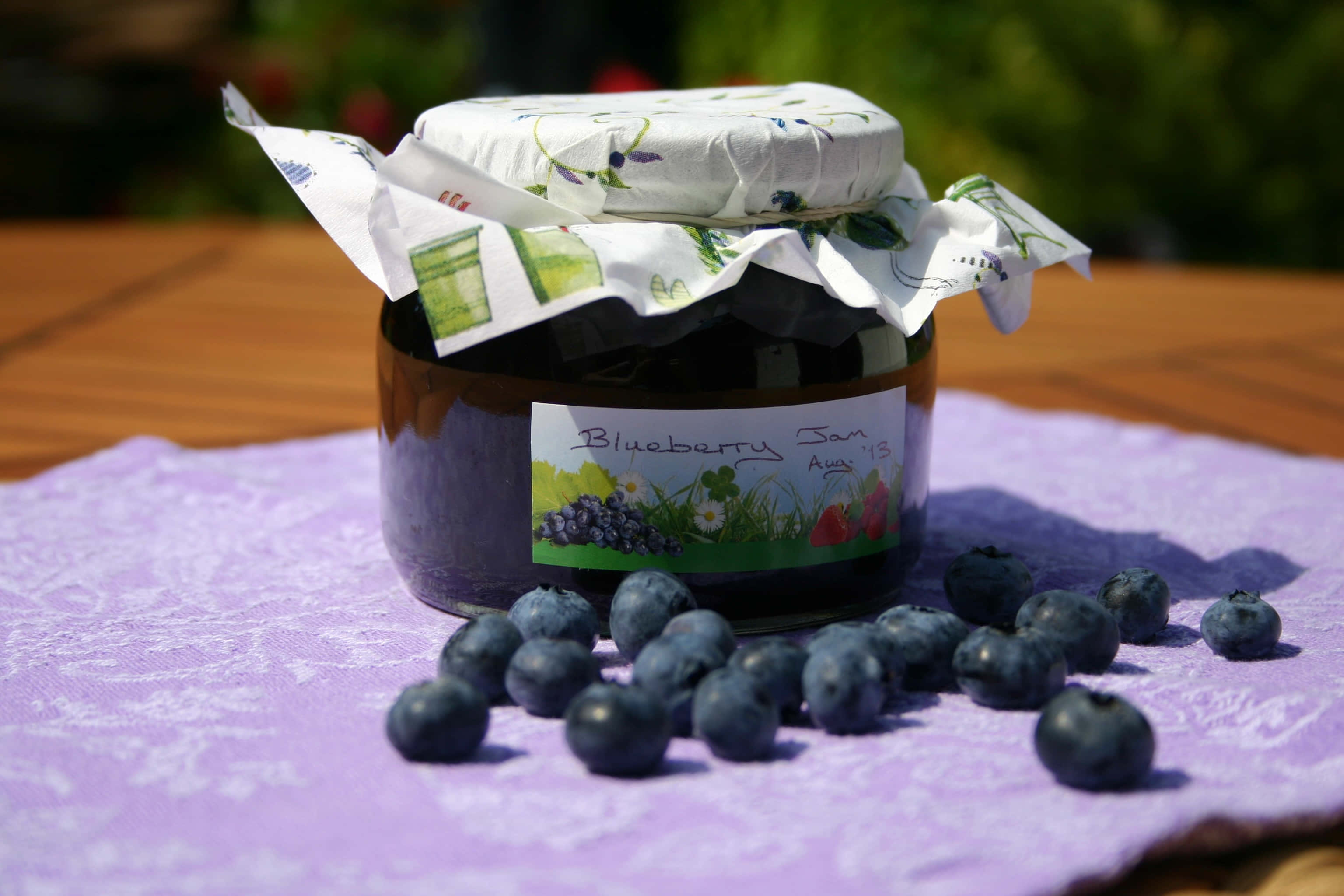 Blueberry Jam 桌布 壁紙 — 一罐自製藍莓醬搭配新鮮莓果，放在紫色布上，在明亮陽光下。