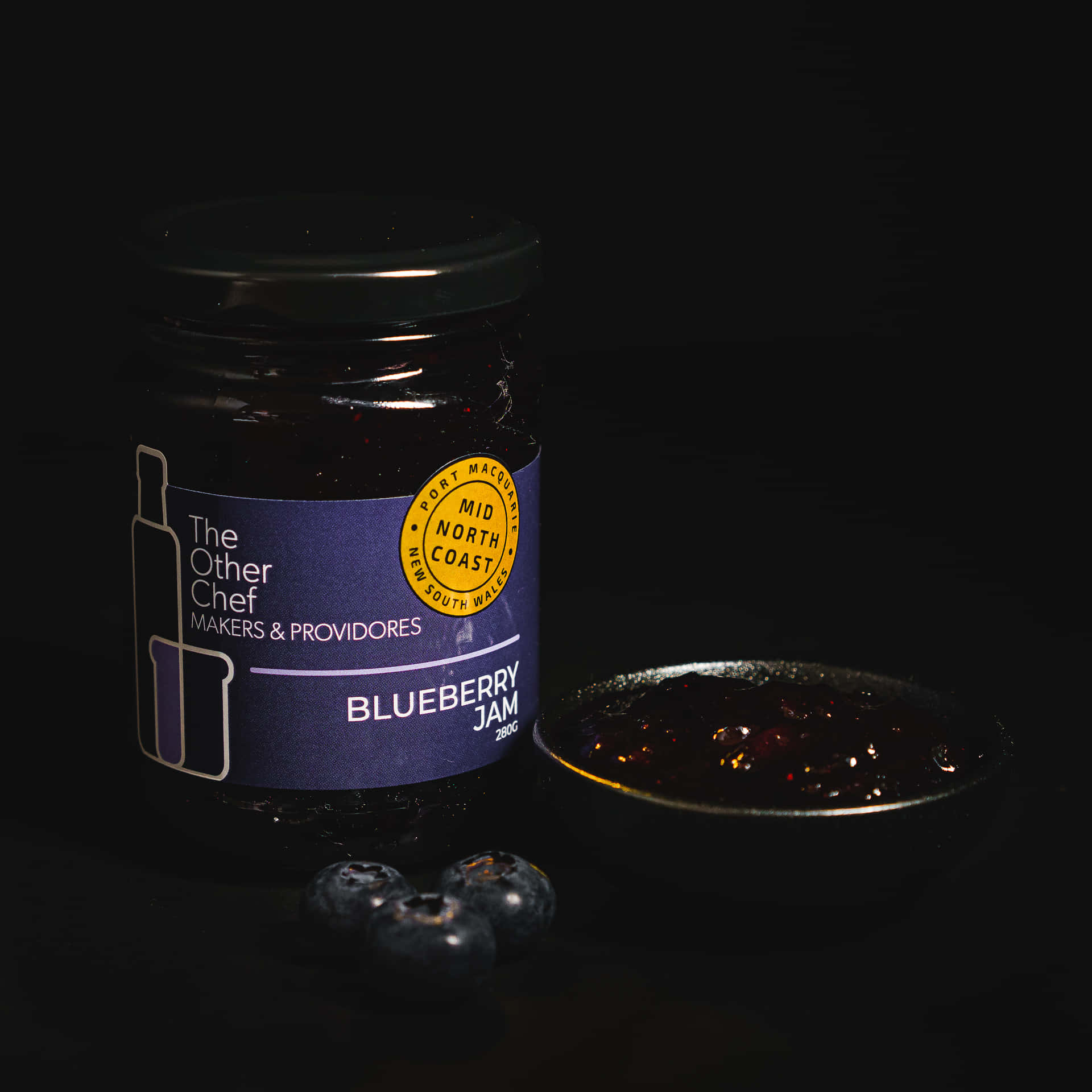 Blueberry Jam 桌布 壁紙 — 一罐藍莓果醬，旁邊有新鮮莓果和一匙果醬，在黑色背景下戲劇性地打光。