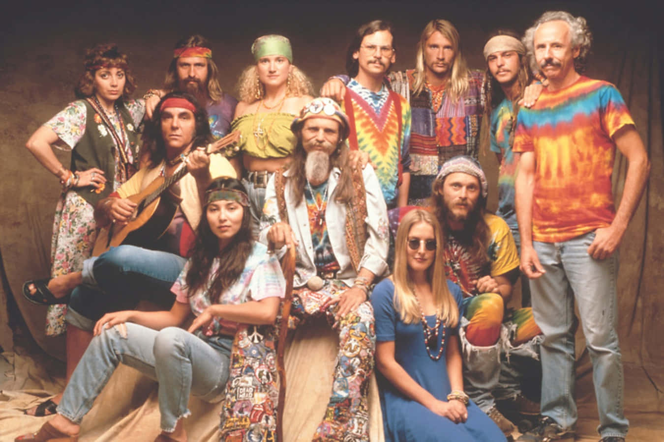 嬉皮 桌布 壁紙 — 一群11人身穿1970年代嬉皮服裝，在素色背景前一起擺姿勢，穿著色彩繽紛的紮染襯衫和頭帶。