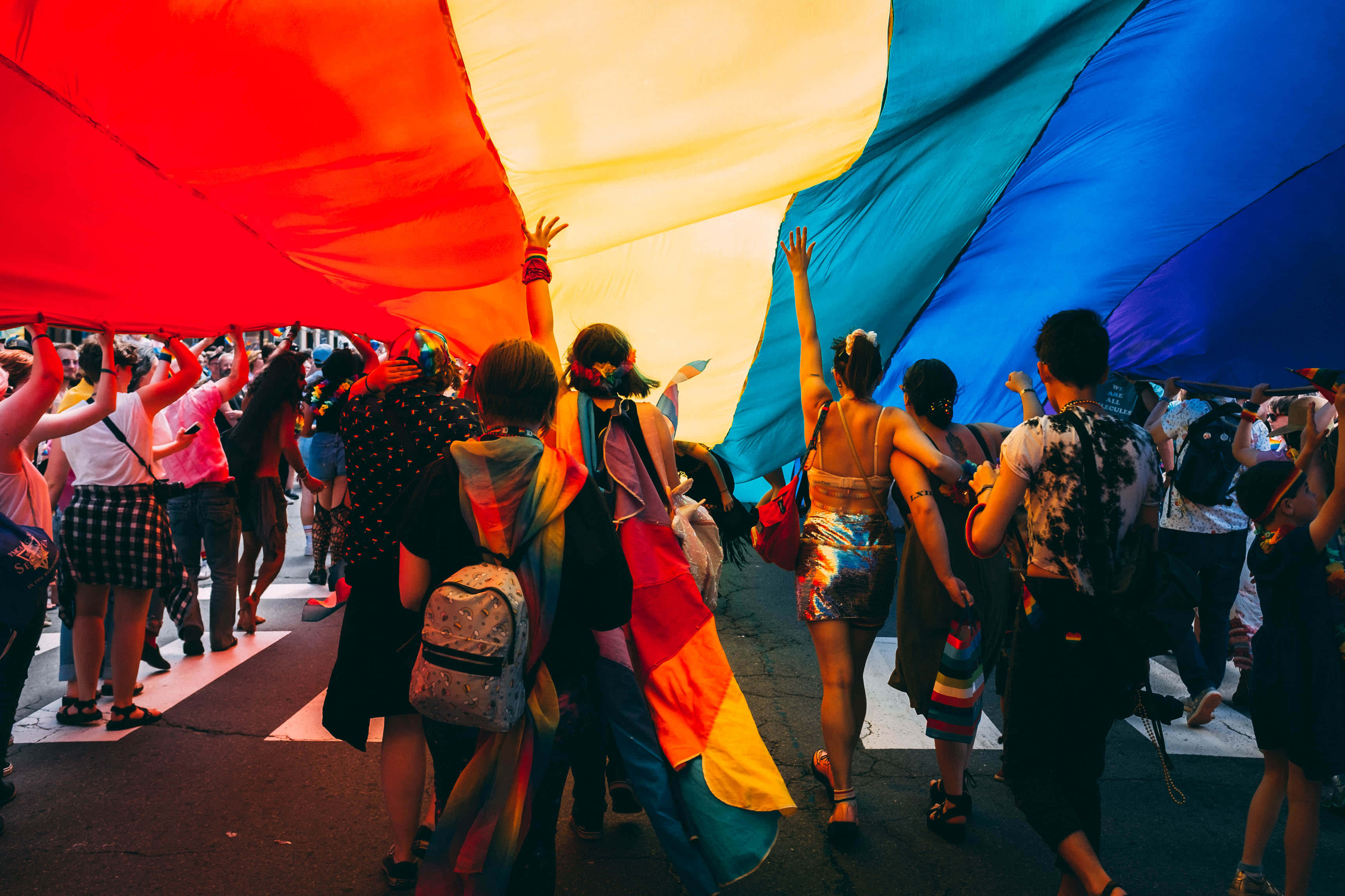 Flag 桌布 壁紙 — 一群不同背景的人沿著街道行走，在溫暖的金色光芒下高舉一面巨大的彩虹旗。