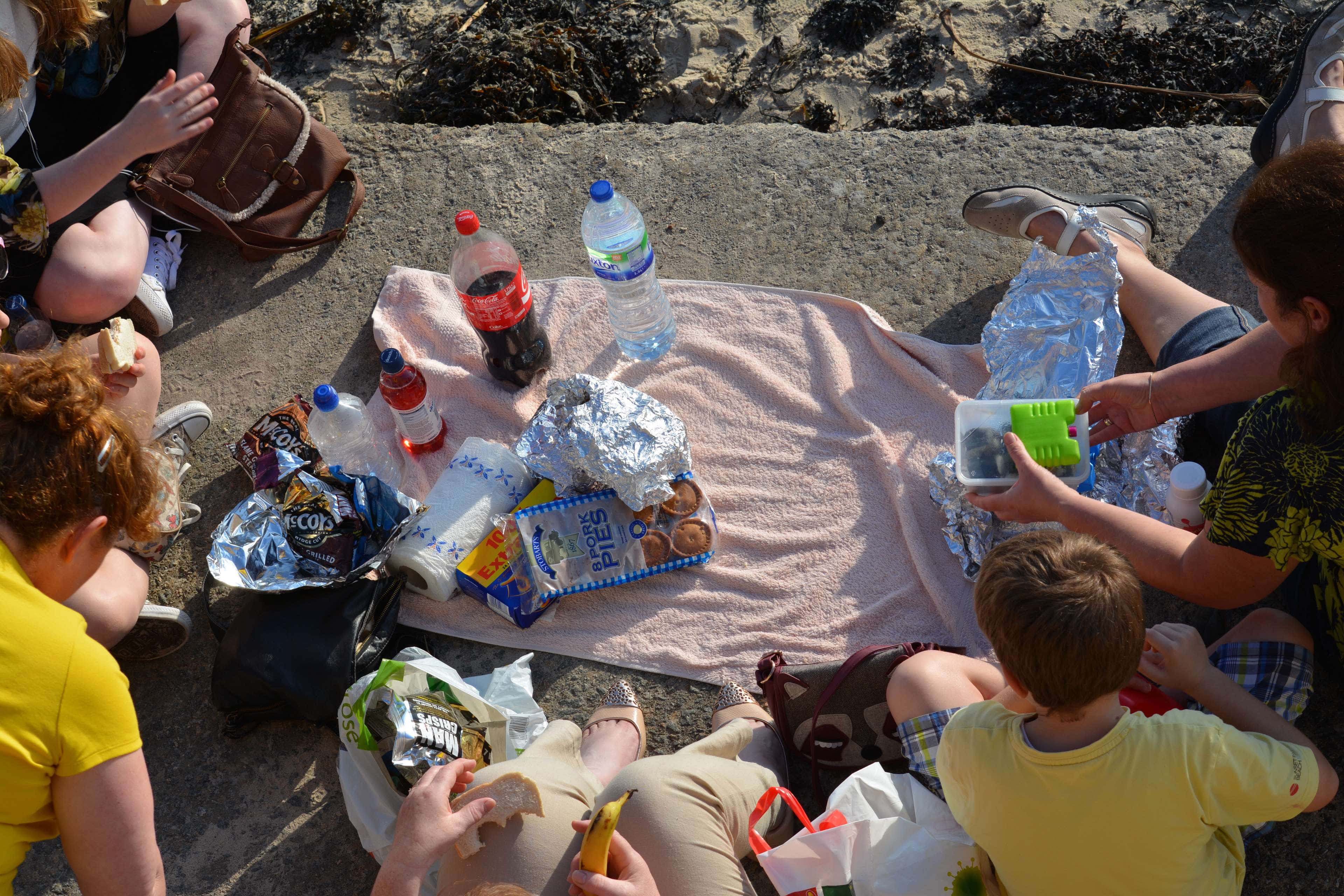 Beach Picnic 桌布 壁紙 — 一群人和小孩在粉紅色毛巾上享受陽光沙灘野餐，有零食、飲料和錫箔紙包裹的食物。