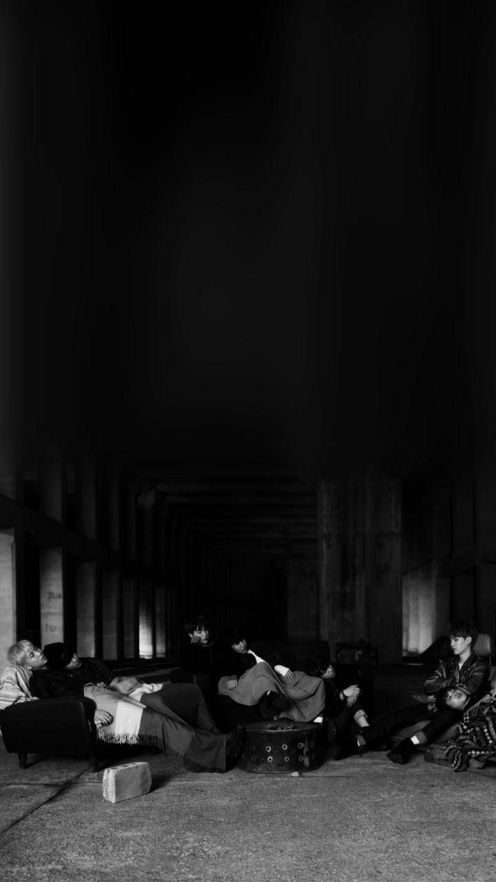 BTS 黑色 桌布 壁紙 — 一群人在昏暗的柱廊走廊中休息，黑白風格，帶有戲劇性的陰影和極少的光源。