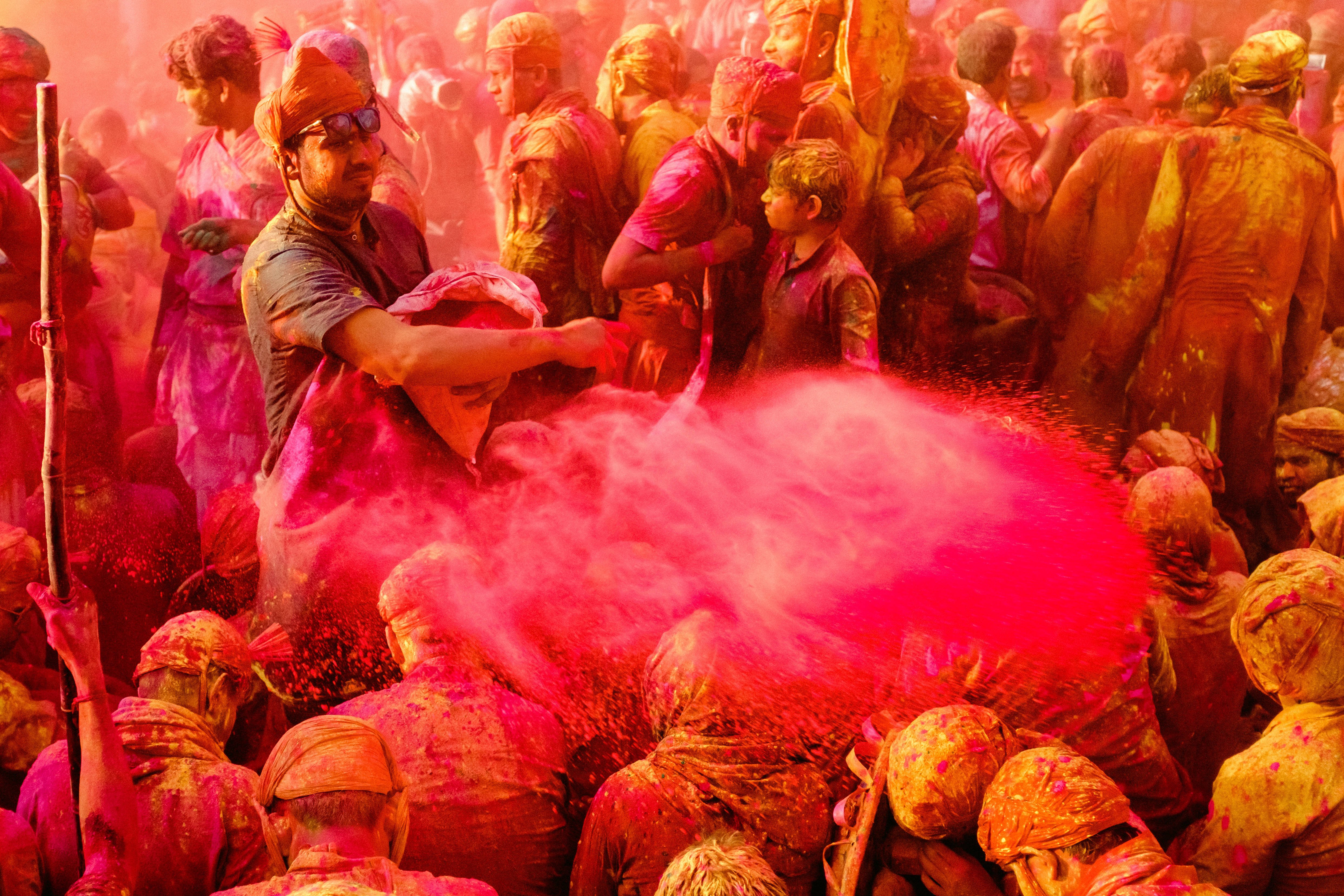 Holi 桌布 壁紙 — 一群人在色彩繽紛的節日中歡快地拋灑鮮豔的粉紅色粉末，人們身上覆蓋著紅色、橙色和粉紅色的色調。