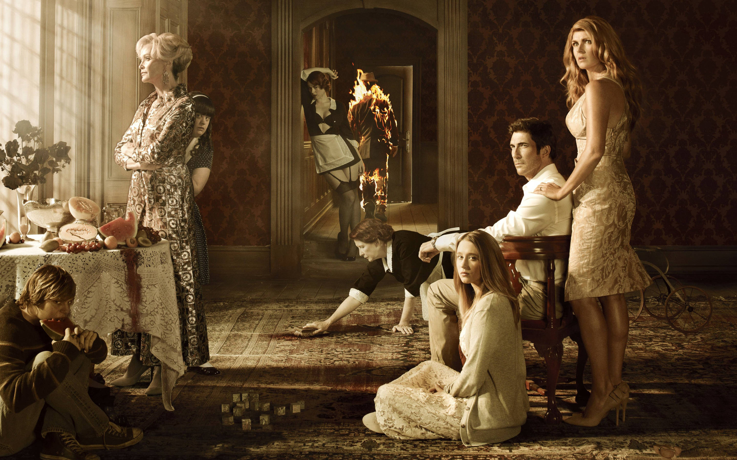 American Horror Story 桌布 壁紙 — 一群人在豪華、昏暗的豪宅房間中，帶有戲劇性的燈光，背景中有一個僕人拿著燃燒的火把。