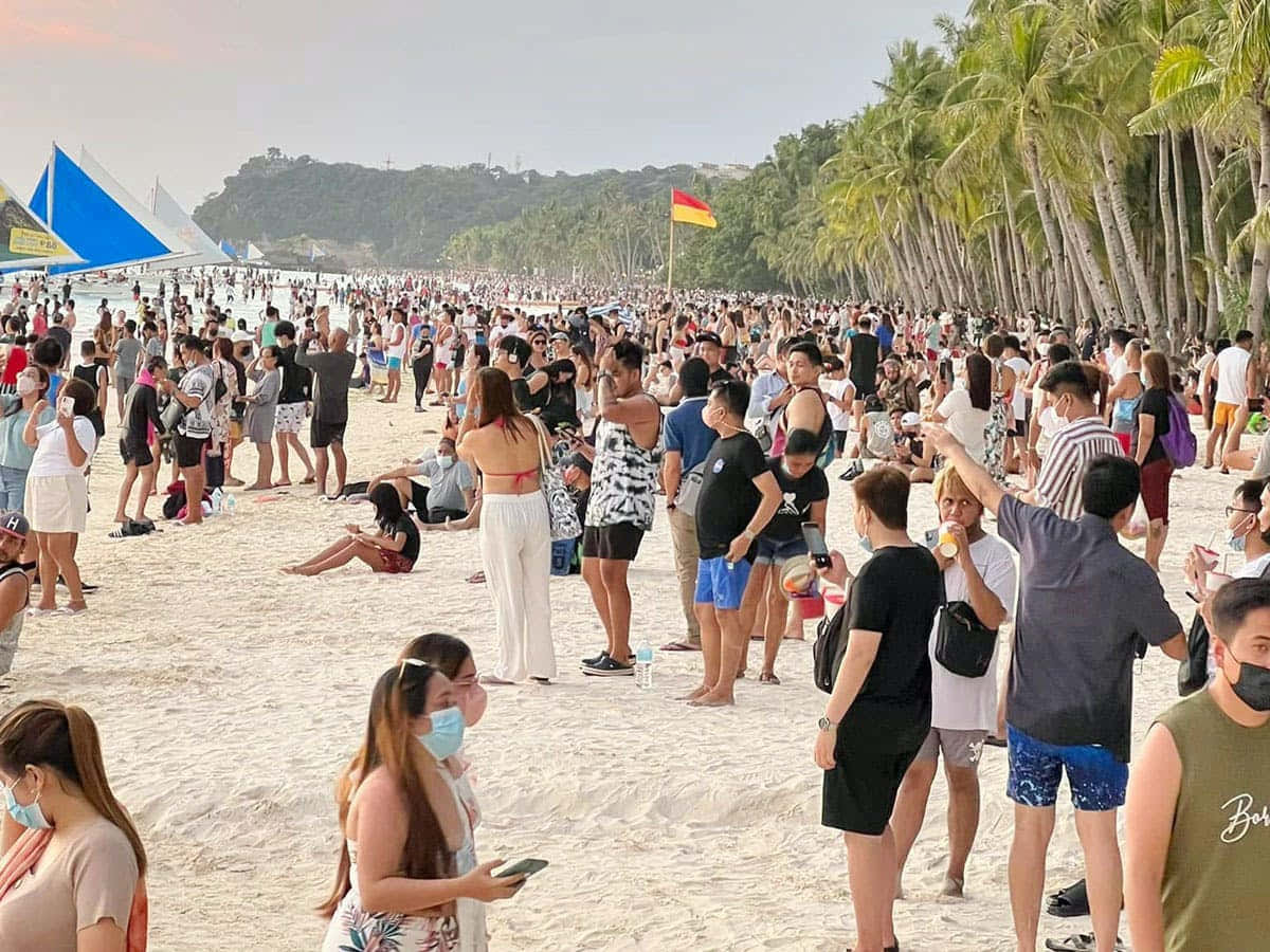 節日 桌布 壁紙 — 一群人在陽光明媚的海灘上戴口罩，遠處有棕櫚樹和帆船。
