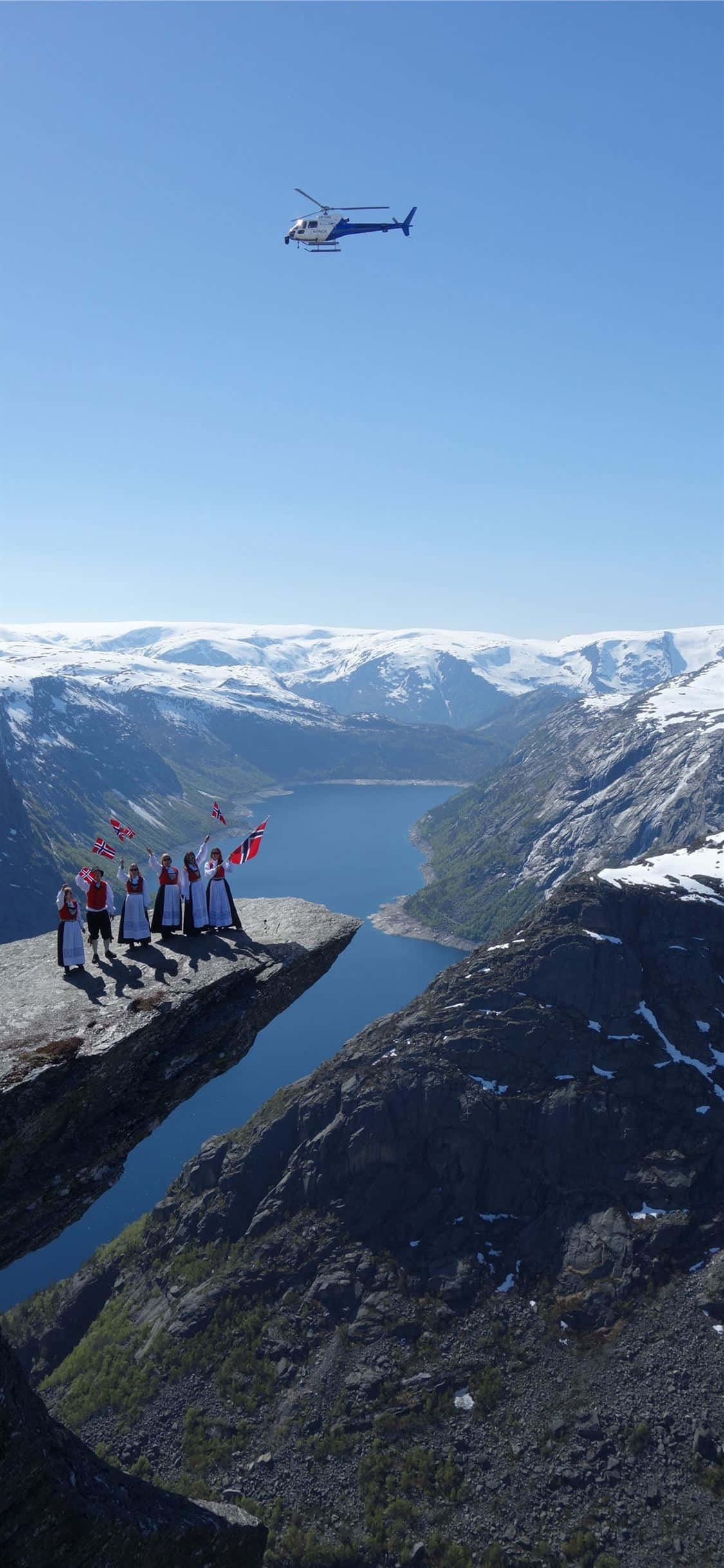 Norway 桌布 壁紙 — 一群人站在懸崖邊緣，俯瞰著深峽灣，峽灣兩旁是白雪覆蓋的山脈，天空清澈湛藍，一架直升機在頭頂飛過。