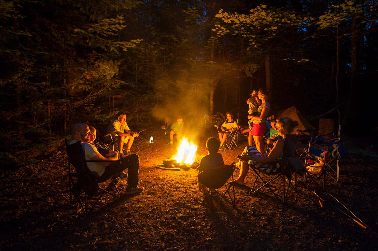 Camping 桌布 壁紙 — 一群人聚集在黑暗森林中閃爍的營火周圍，夜晚被溫暖的橙色火焰照亮。