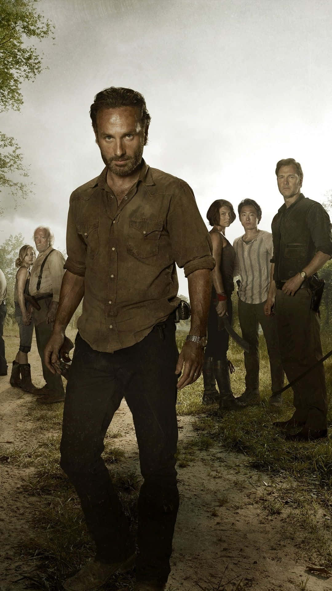 The Walking Dead iPhone 桌布 壁紙 — 一群倖存者沿著一條泥土小徑穿過雜草叢生的荒野，天空多雲。