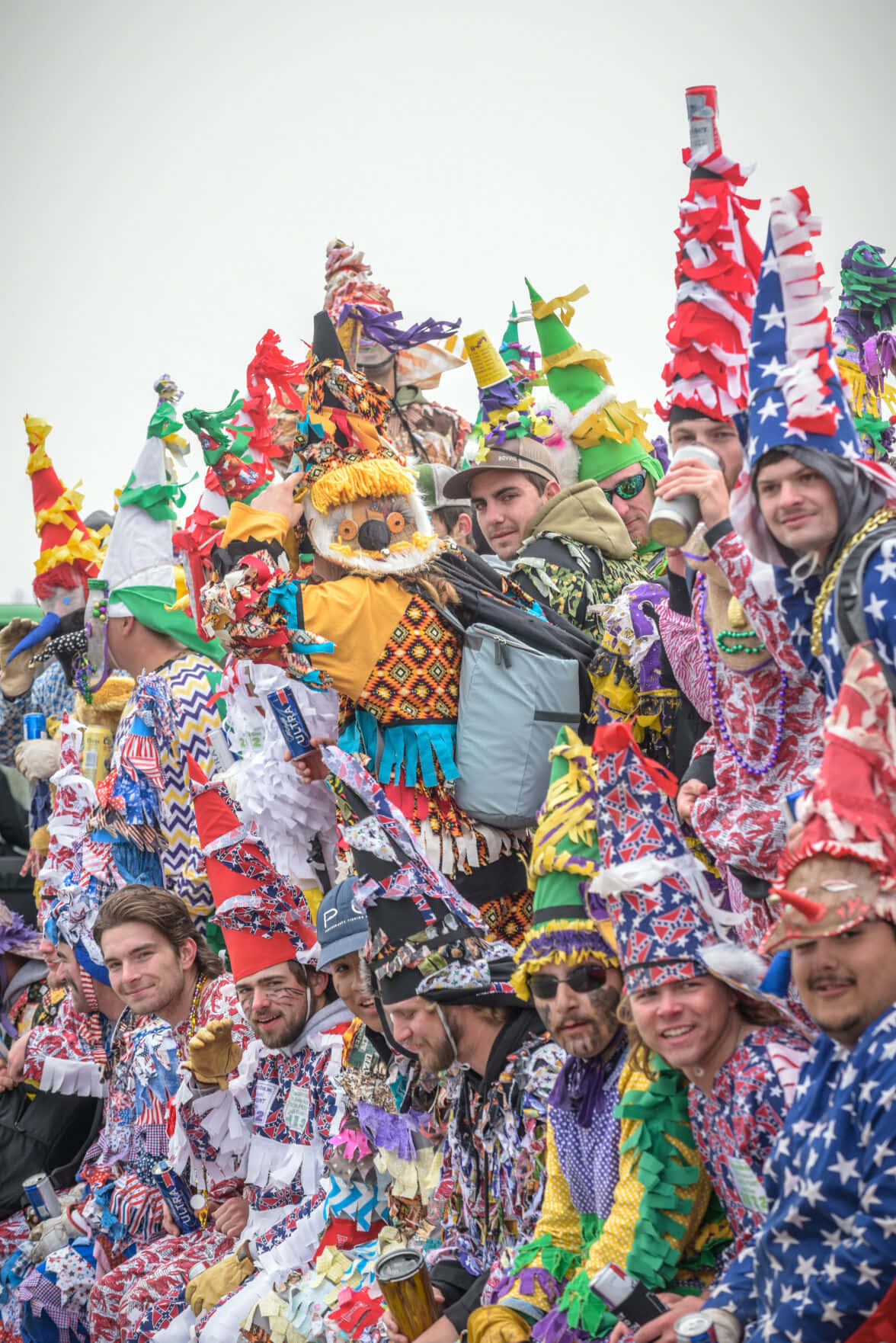 Mardi Gras 桌布 壁紙 — 一群充滿活力的人穿著色彩繽紛的手工服裝和高帽子，在多雲的天空下於熱鬧的節日中微笑慶祝。