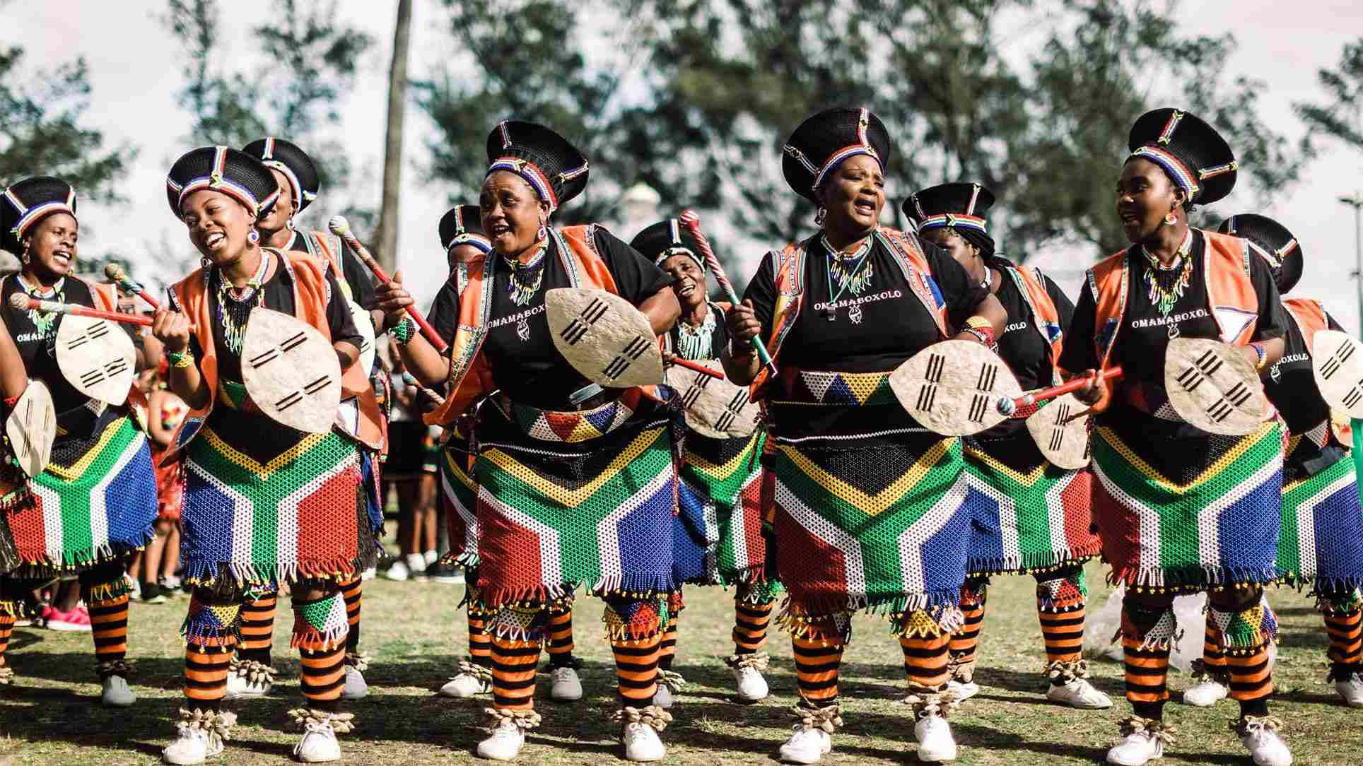 African Culture 桌布 壁紙 — 一群充滿活力的女性穿著色彩繽紛的傳統服飾，在明亮的日光下帶著手鼓歡快地戶外跳舞。