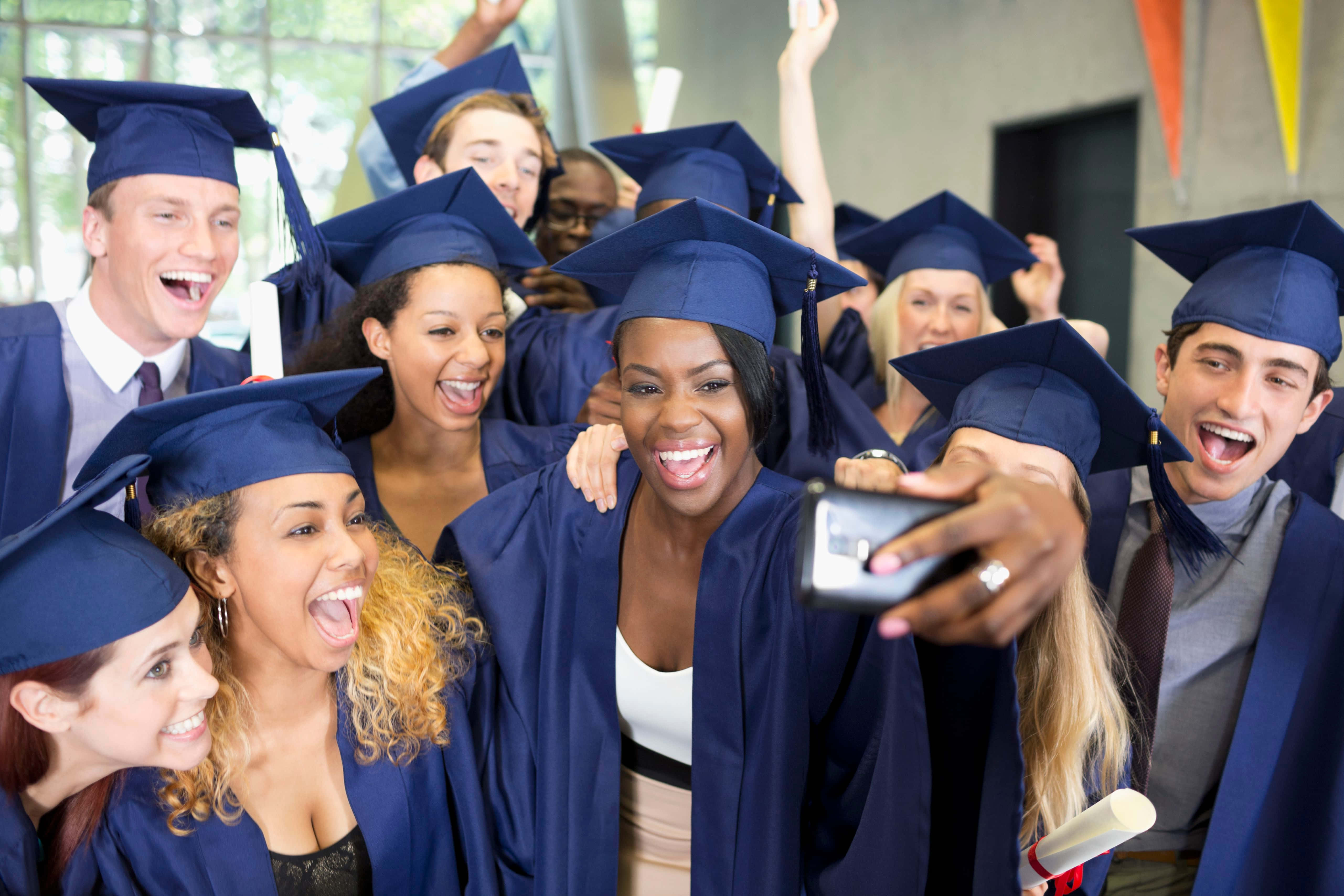 Graduation 桌布 壁紙 — 一群多元化的畢業生，身穿藍色學士帽和袍，在室內愉快地一起自拍。