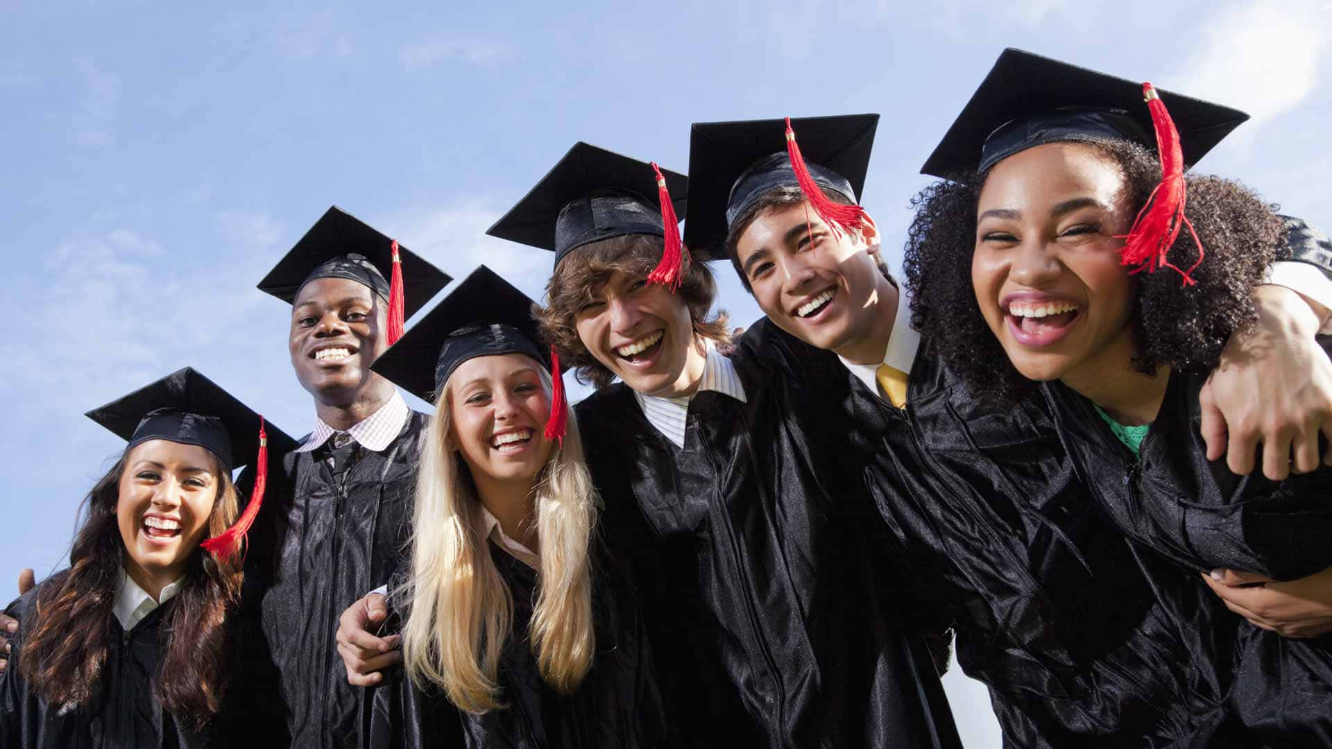 Graduation 桌布 壁紙 — 一群多元化的畢業生，身穿黑色學士帽和學士袍，在明亮的藍天下一起開懷大笑。