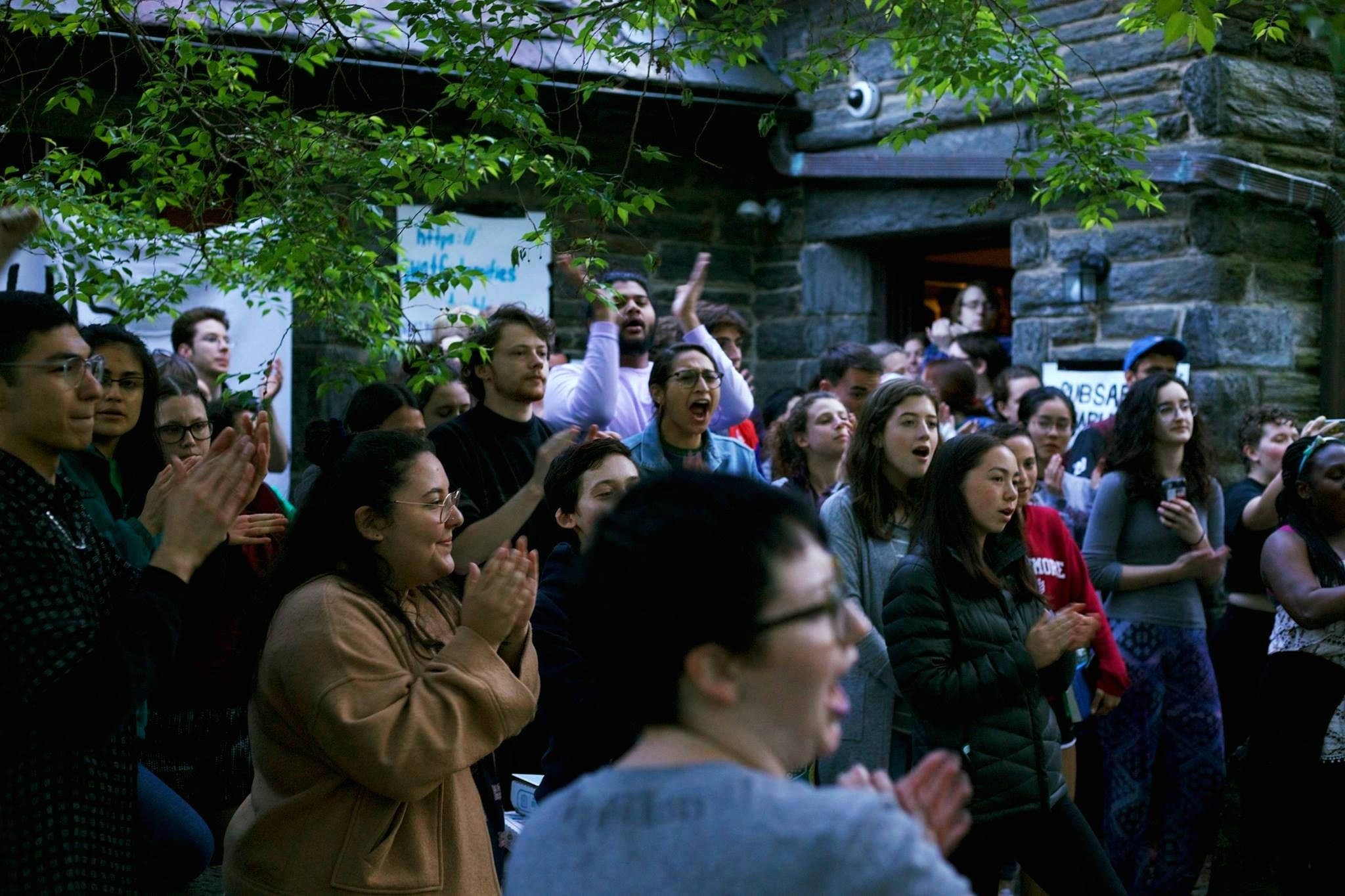 college 桌布 壁紙 — 一群多元化的群眾在綠樹成蔭的石建築外熱情歡呼鼓掌。