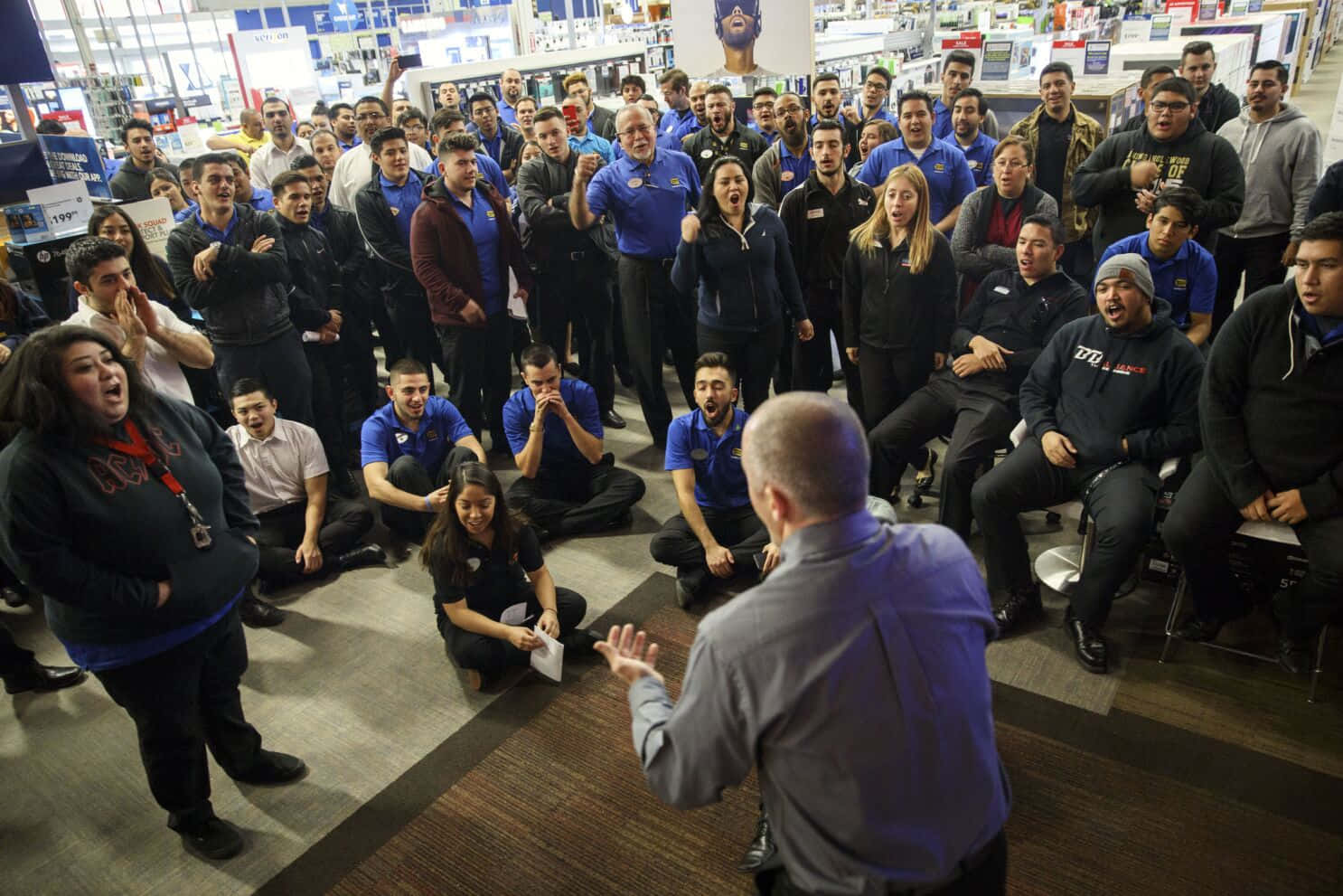 Best Buy 桌布 壁紙 — 一群多元化的零售店員工在一個大型開放空間中熱情地歡呼並對演講者作出反應。