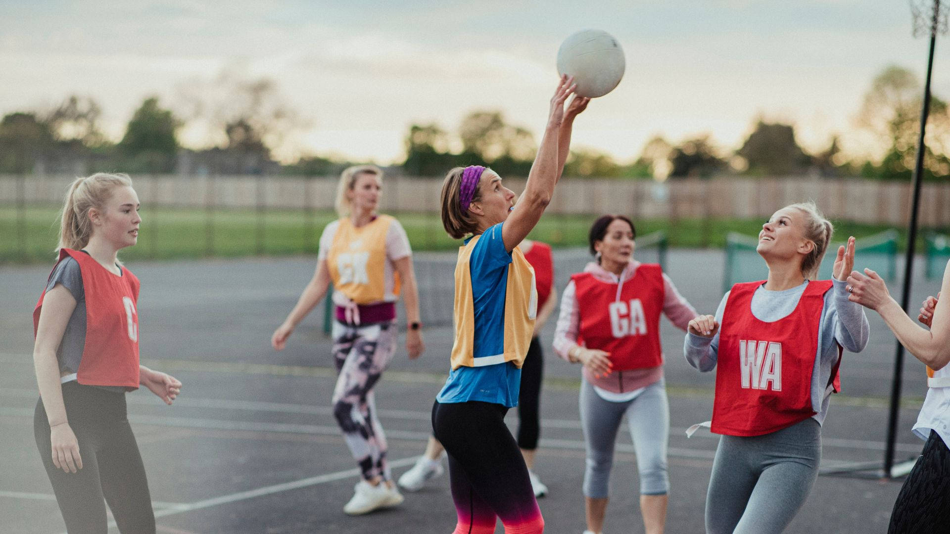 ball 桌布 壁紙 — 一群女性在日落時分的戶外球場上打籃網球，其中一名球員跳起接球。