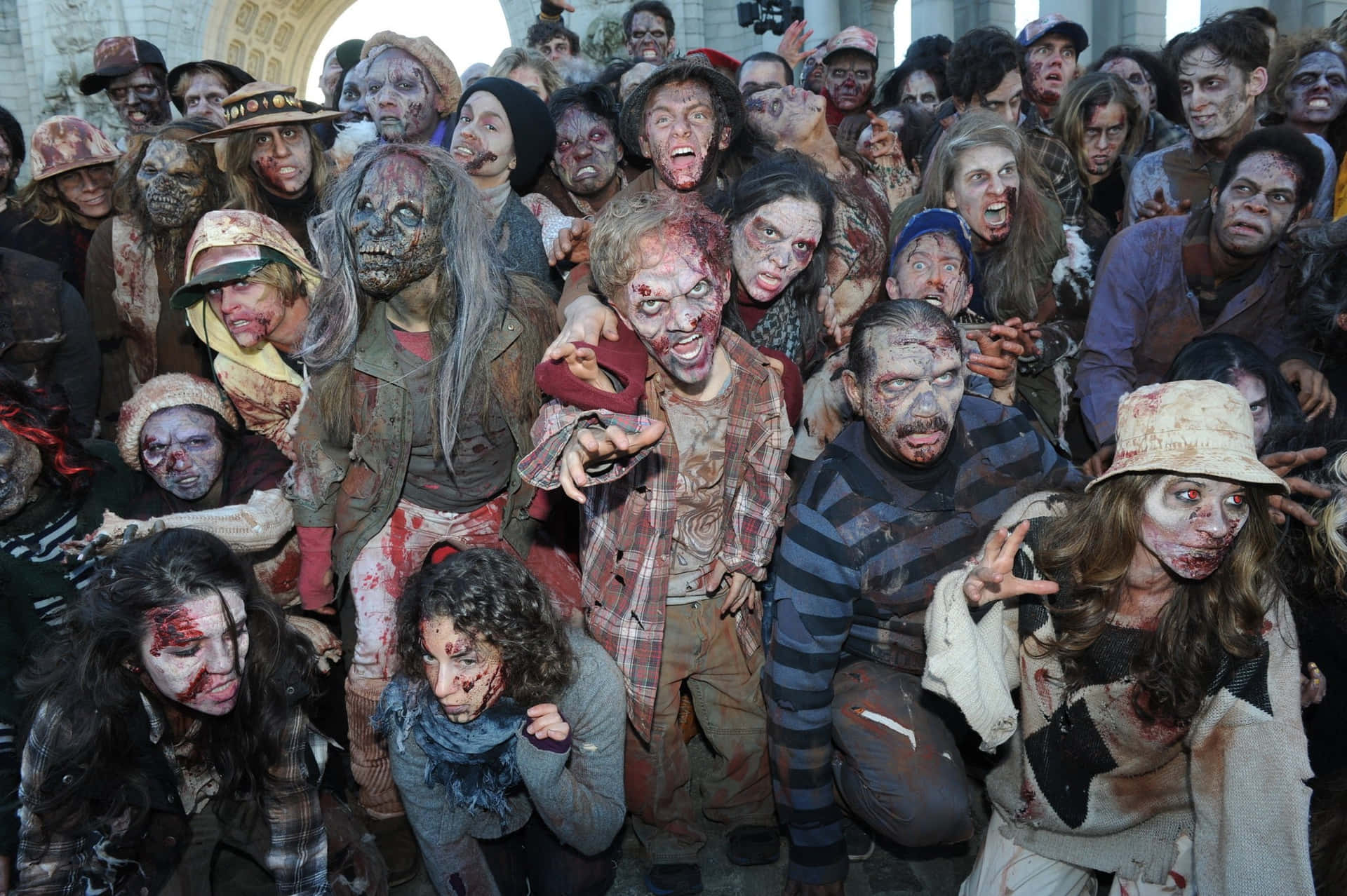 Zombie 桌布 壁紙 — 一群密集的怪誕、血淋淋的殭屍，穿著破爛衣服，帶著驚恐的表情，在混亂恐怖的場景中蜂擁前進。