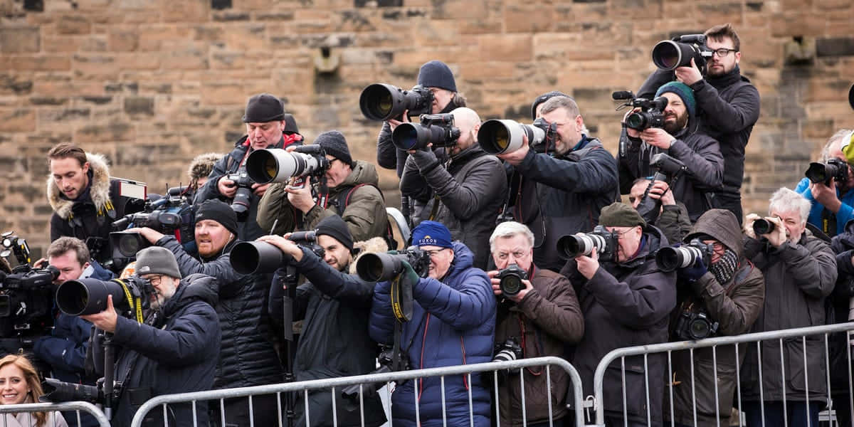 Paparazzi 桌布 壁紙 — 一群密集的攝影師帶著大型望遠鏡頭，在石牆前的金屬路障後捕捉瞬間。