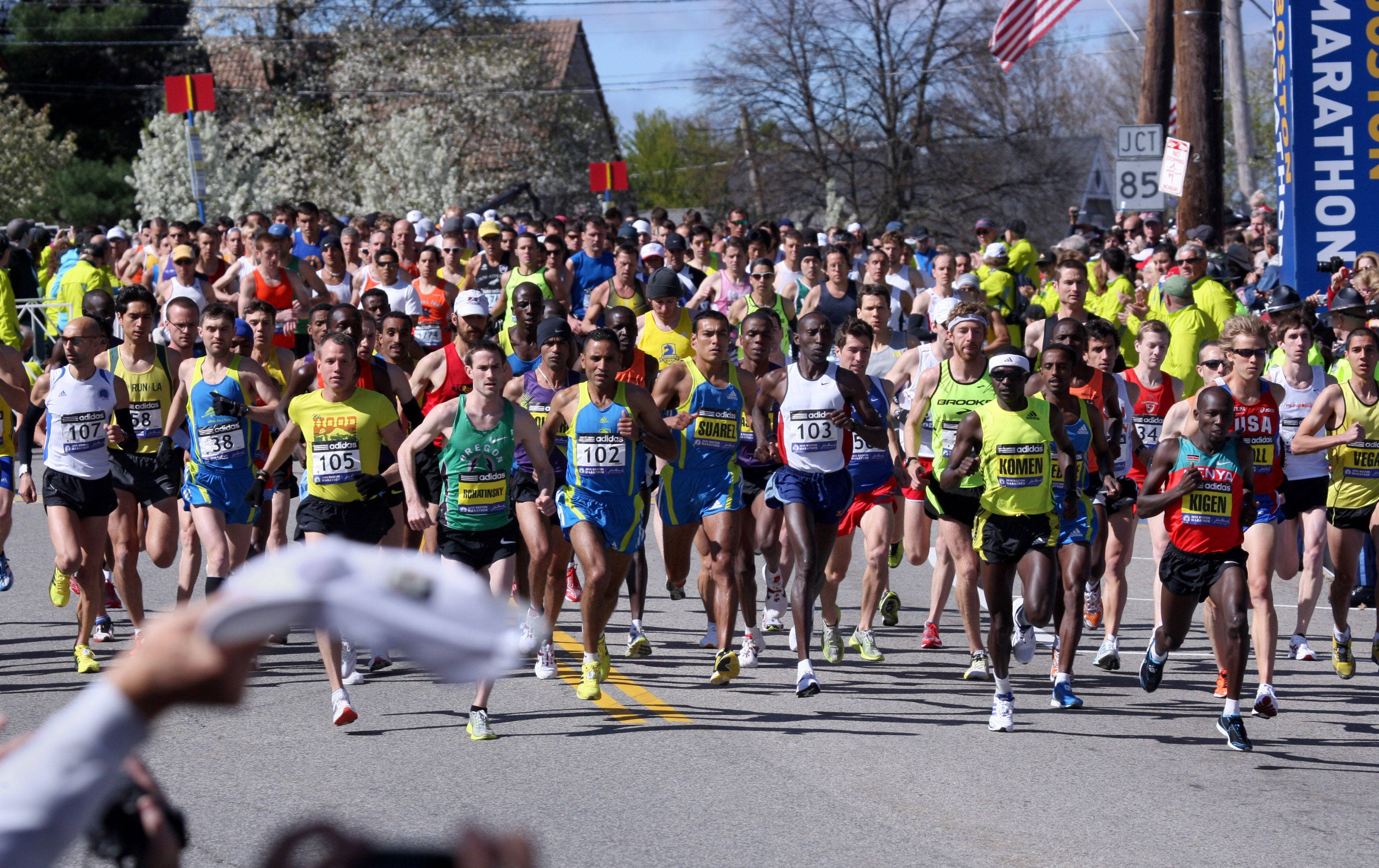 Marathon 桌布 壁紙 — 一群密集的馬拉松跑者穿著色彩繽紛的運動裝備，在陽光明媚的街道上衝刺，街道兩旁擠滿觀眾和春天的花朵。
