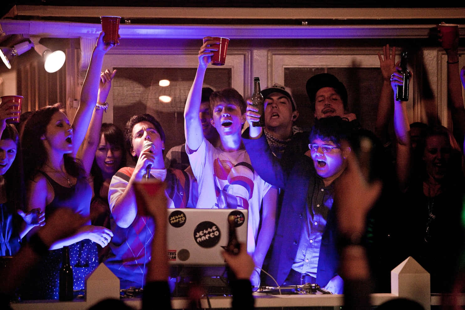House Party 桌布 壁紙 — 一群年輕人在夜店中瘋狂歡呼，紫色燈光與高舉的紅色杯子。