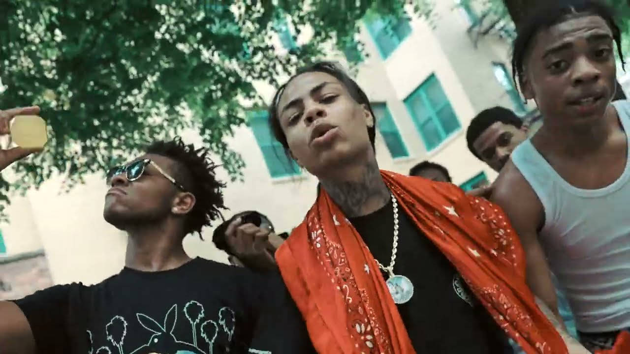 Kay Flock 桌布 壁紙 — 一群年輕男子在戶外擺姿勢，其中一人手持金幣，位於大樓附近綠樹成蔭的樹下。