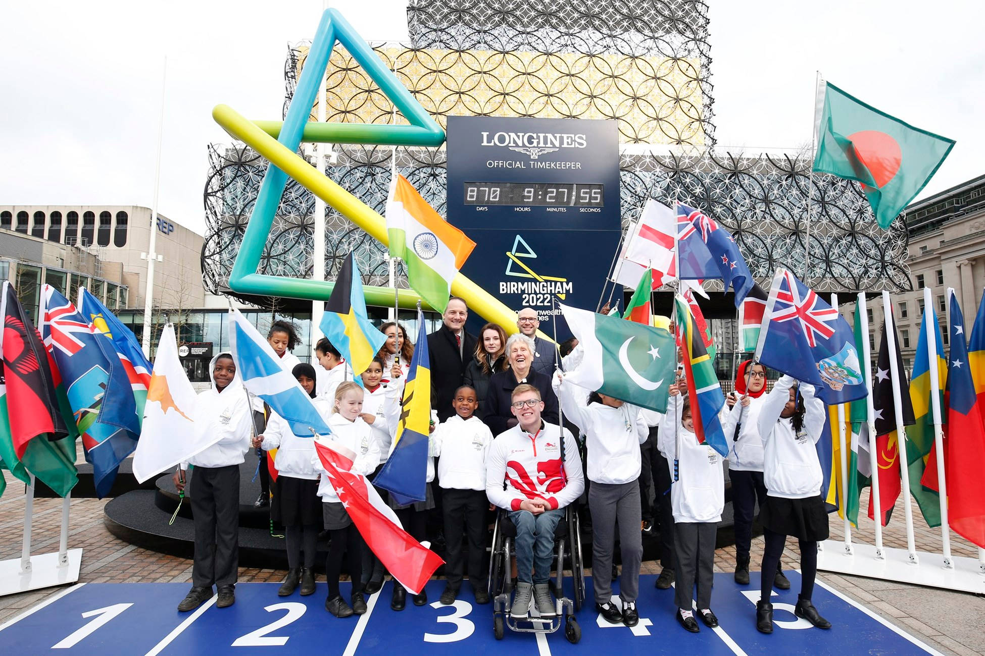 Athlete 桌布 壁紙 — 一群年輕運動員和官員在伯明罕2022大英國協運動會上，自豪地手持國旗，圍繞著一個大型浪琴計時顯示屏合影。