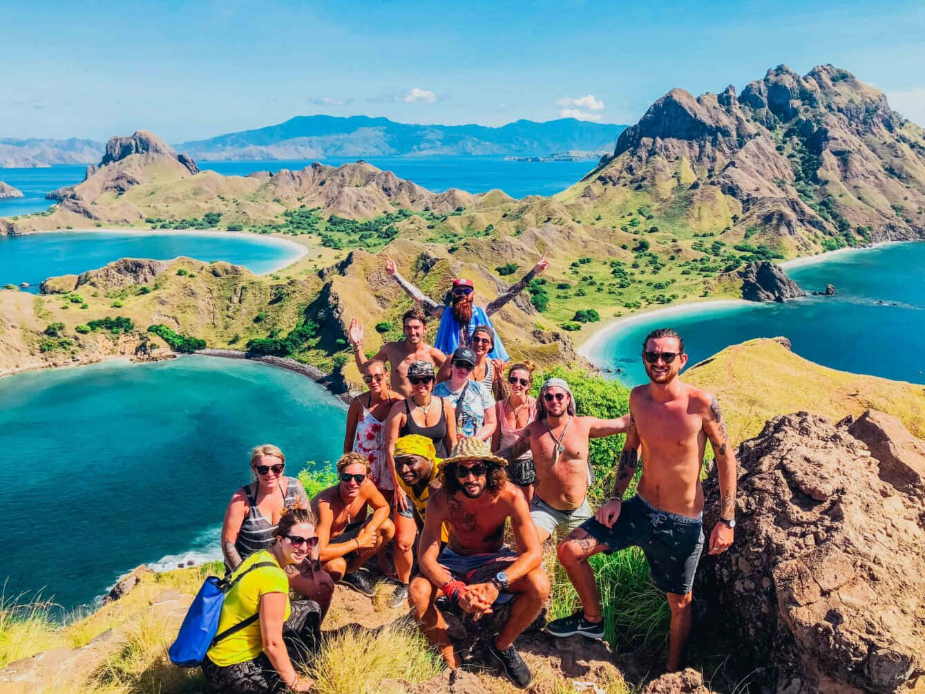 島嶼 桌布 壁紙 — 一群微笑的旅行者在長滿青草的懸崖上擺姿勢，俯瞰著碧綠的潟湖和崎嶇的綠色山脈，天空清澈湛藍。