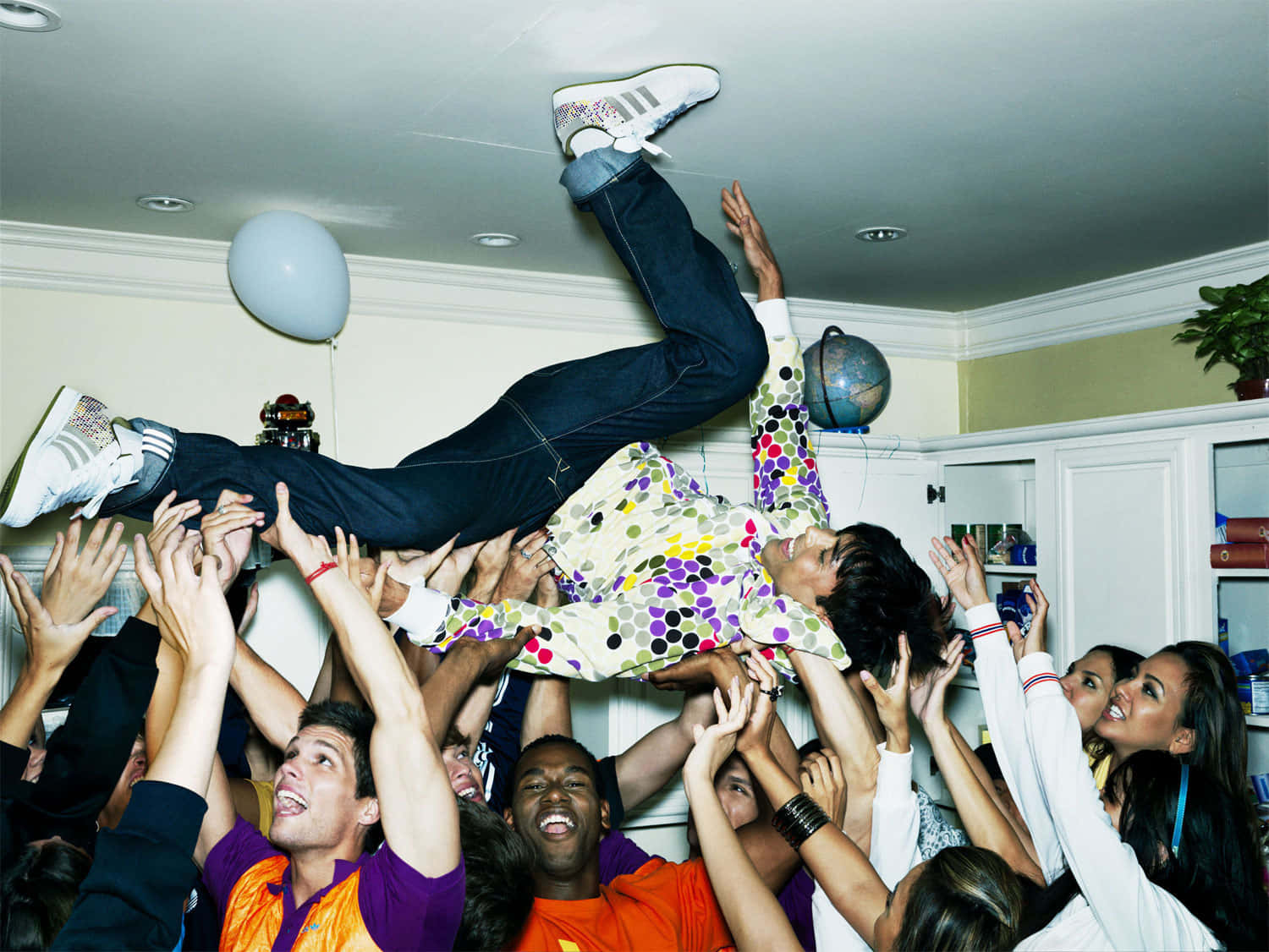 House Party 桌布 壁紙 — 一群快樂的朋友在室內熱鬧的慶祝活動中，將一個人高高舉起，周圍有氣球和鮮豔的衣服。
