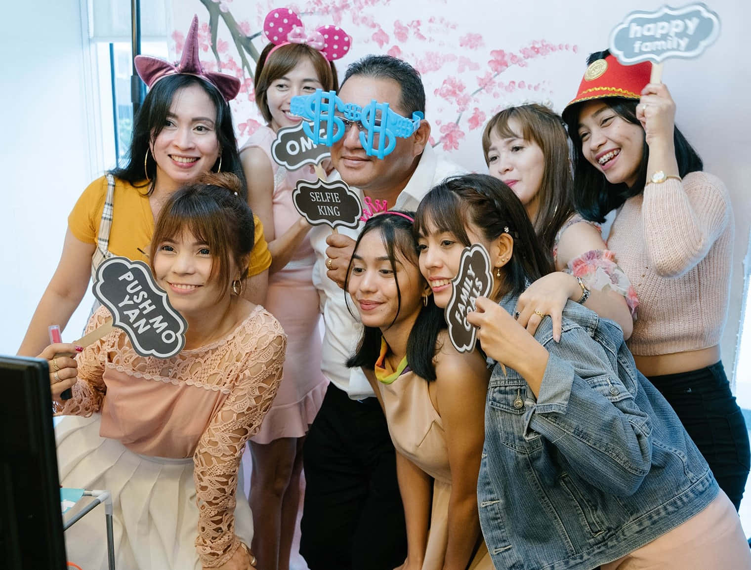 Photo Booth 桌布 壁紙 — 一群快樂的朋友在明亮的室內環境中，用有趣的道具和櫻花背景擺姿勢。