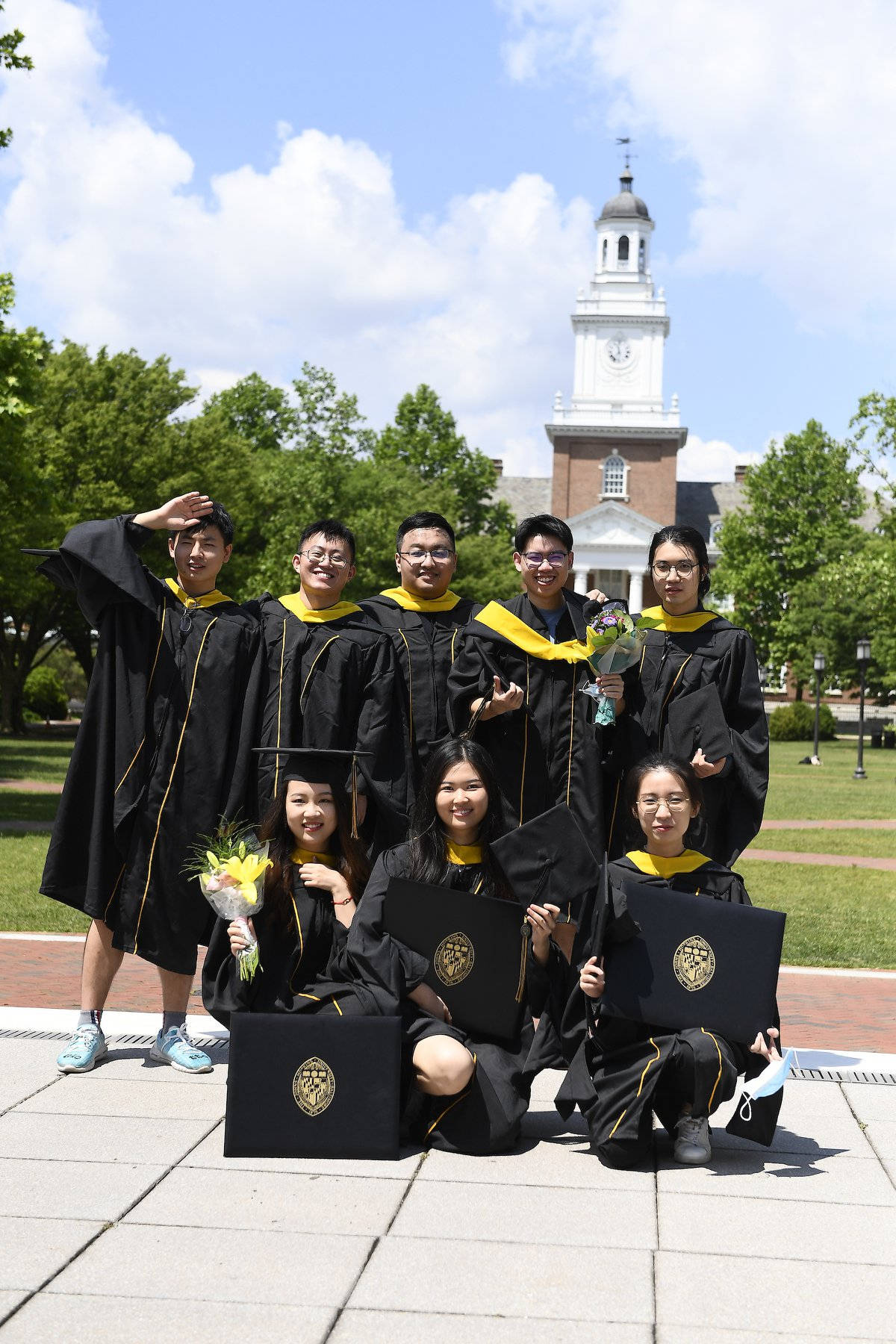college 桌布 壁紙 — 一群快樂的畢業生身穿黑色和金色的畢業袍和帽子，在陽光明媚的校園日，手持畢業證書和鮮花，自豪地在歷史悠久的鐘樓前合影。