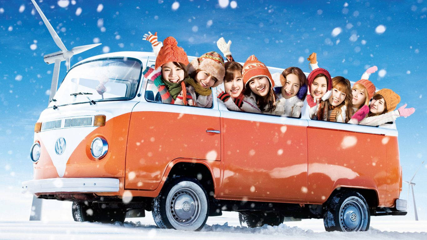 girls 桌布 壁紙 — 一群戴著冬帽的開朗年輕女性，在雪花飄落、藍天白雲和風力發電機的背景下，從一輛復古橙色和白色的VW廂型車中揮手。