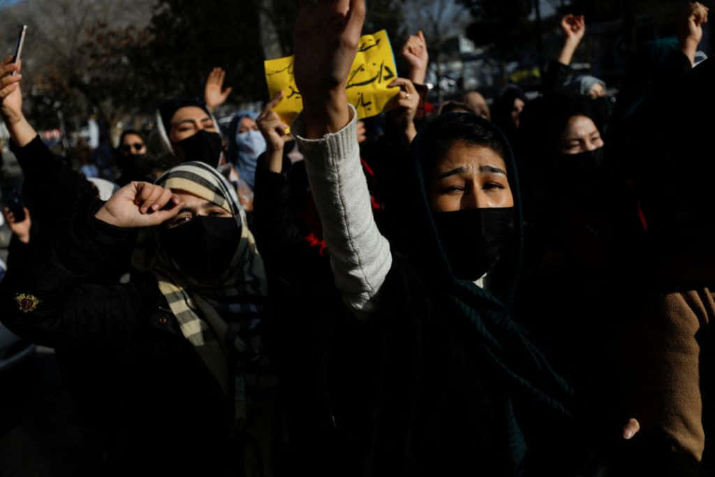 Taliban 桌布 壁紙 — 一群戴著口罩的抗議者在昏暗的傍晚光線下於城市環境中高舉拳頭和標語牌。