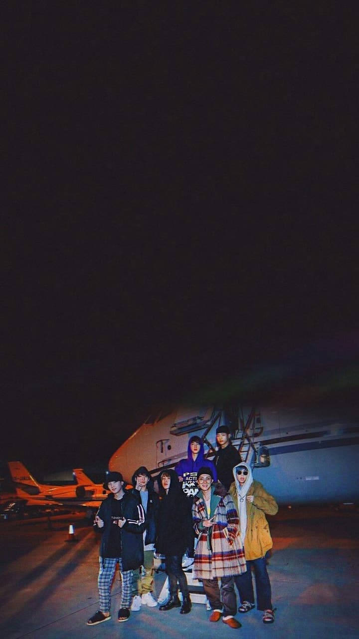 BTS 黑色 桌布 壁紙 — 一群朋友在夜晚的機場停機坪上合照，背景是亮著燈的飛機。