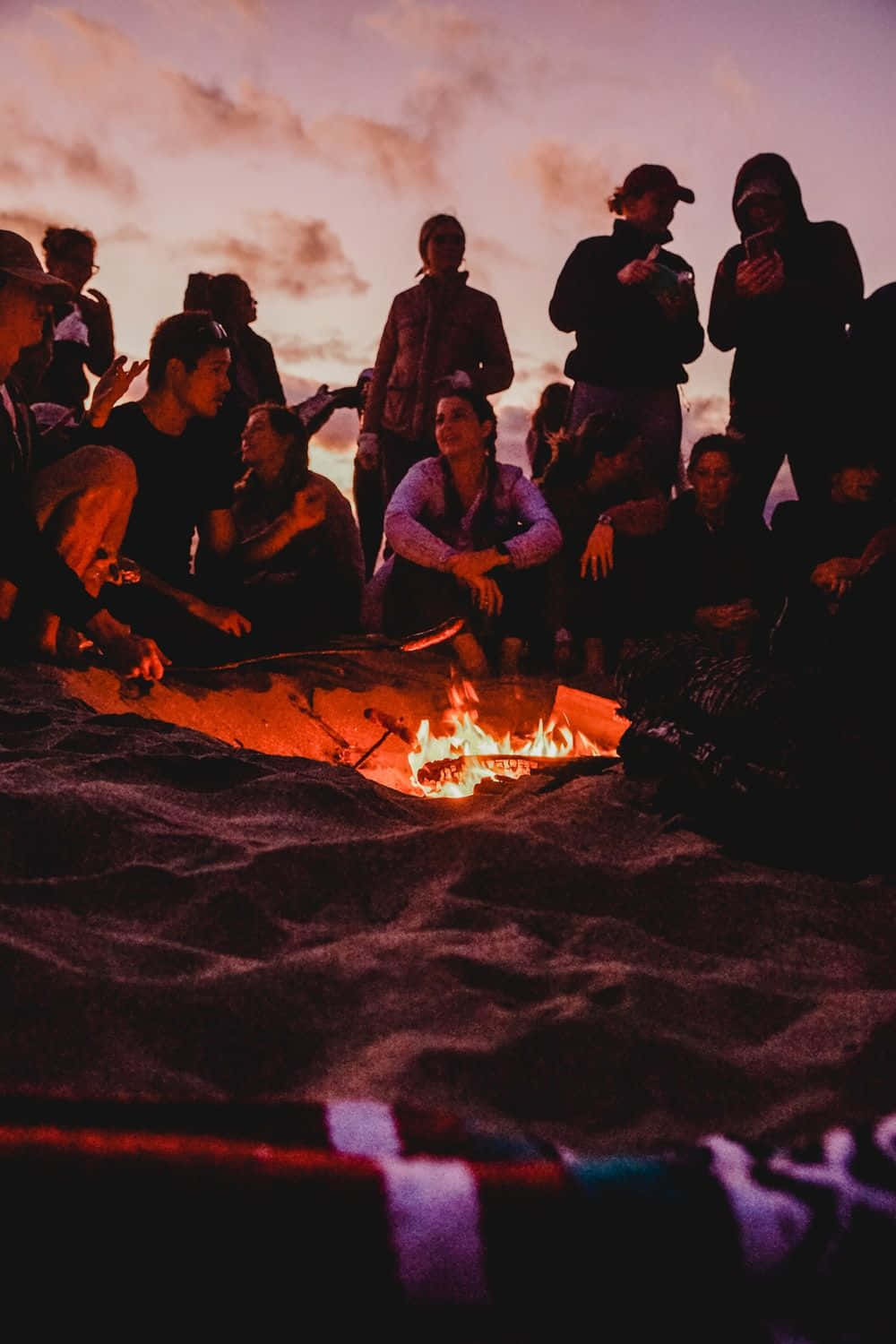 Beach Bonfire 桌布 壁紙 — 一群朋友在黃昏時分聚集在沙灘上發光的營火周圍，在柔和的紫色和橙色天空映襯下形成剪影。