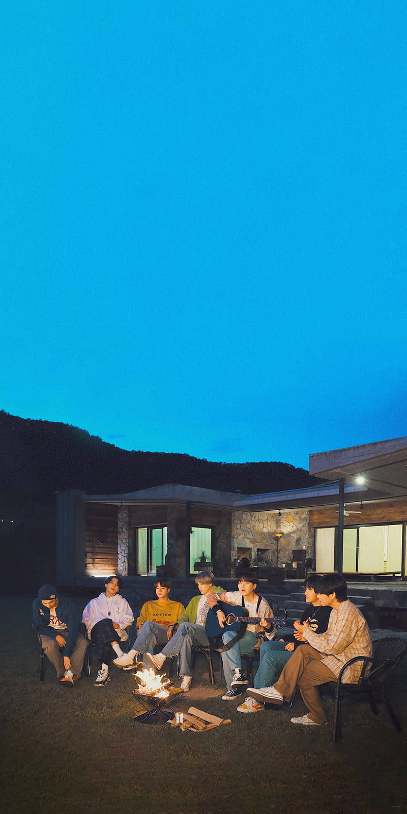 BTS 桌布 壁紙 — 一群朋友在黃昏時分聚集在現代房屋前的營火周圍，背景有山脈。