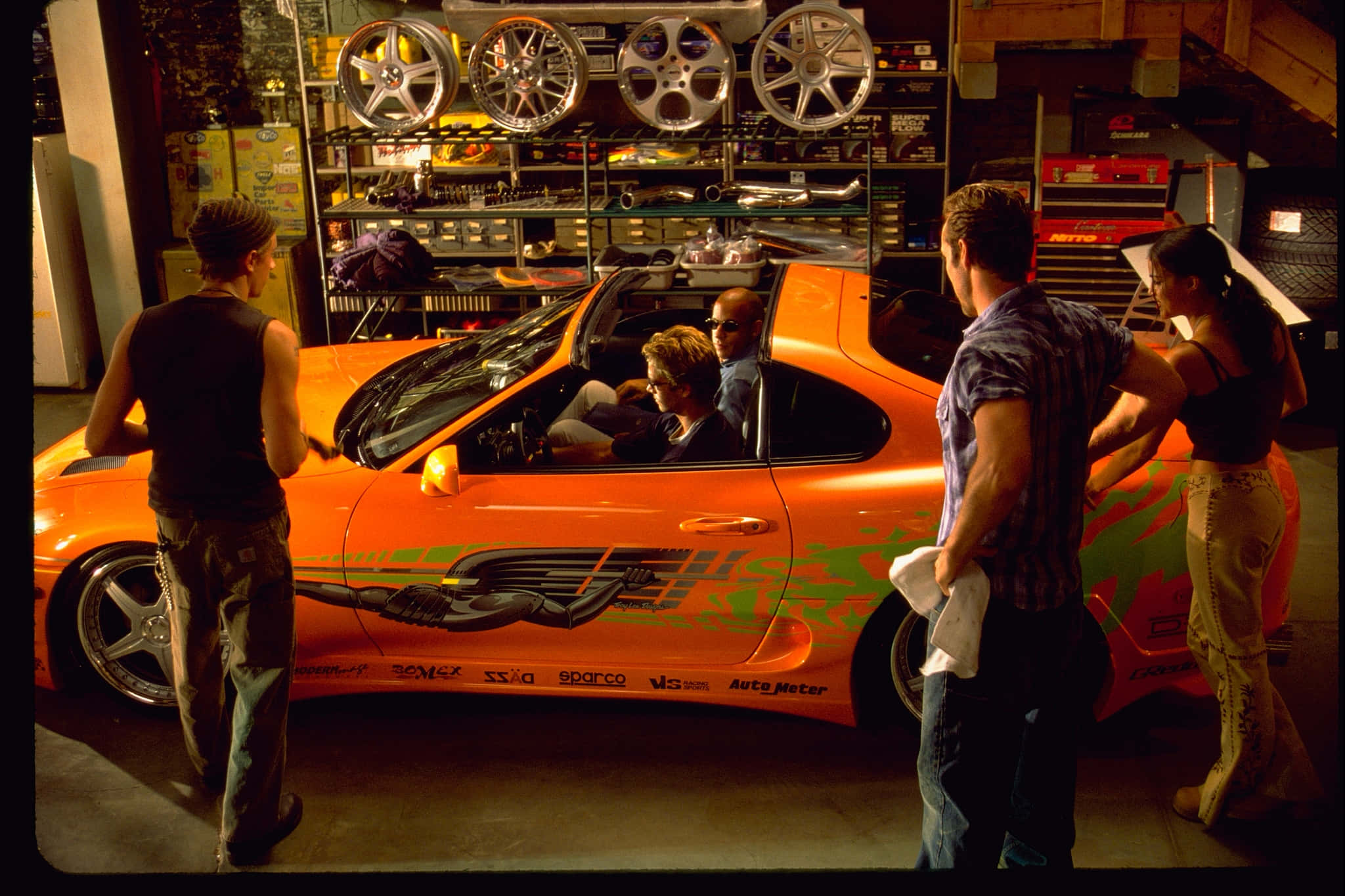 Fast And Furious 1 桌布 壁紙 — 一群朋友聚集在一輛充滿活力的橙色賽車周圍，車庫雜亂，充滿了輪胎和工具。