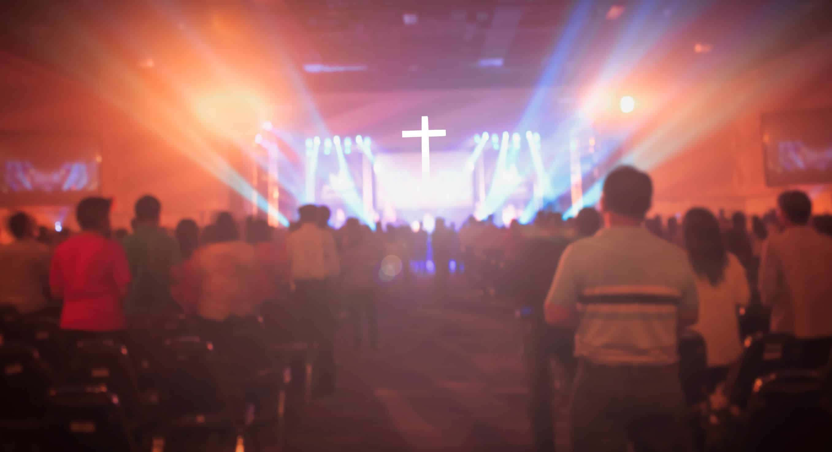 Church Stage 桌布 壁紙 — 一群模糊的人群面向一個明亮的舞台，舞台上有一個發光的十字架和色彩繽紛的聚光燈，在一個敬拜場所。