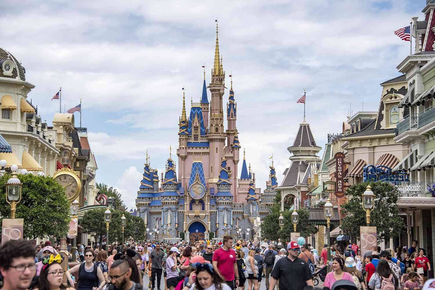 Cinderella 桌布 壁紙 — 一群歡樂的遊客漫步在一條充滿活力的主題公園街道上，通往一座宏偉的粉藍色城堡，天空部分多雲。