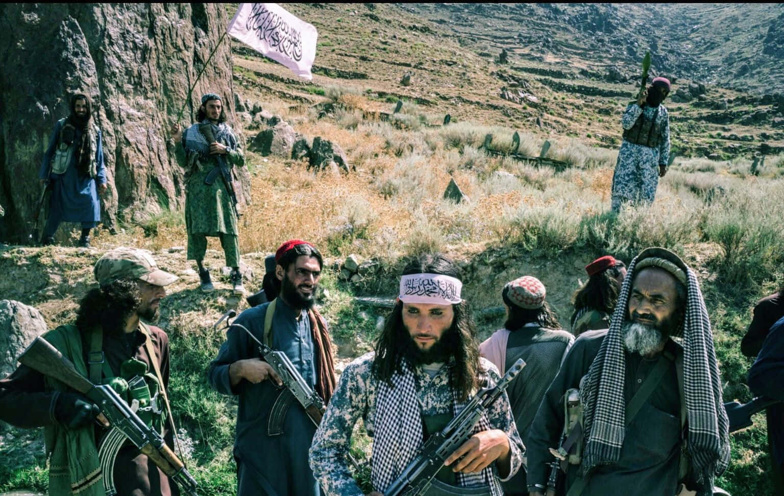 Taliban 桌布 壁紙 — 一群武裝男子在崎嶇的山地地形中，手持步槍並在明亮的日光下揮舞旗幟。