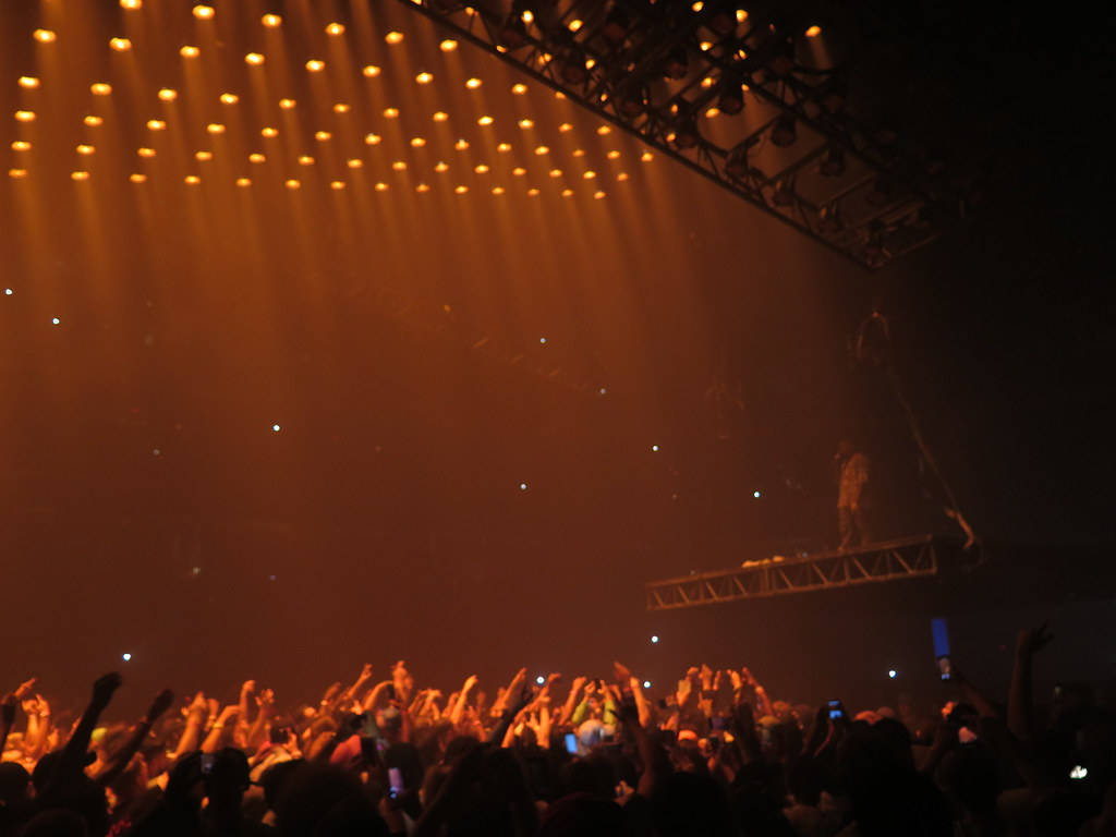 Kanye 桌布 壁紙 — 一群演唱會觀眾在煙霧瀰漫的昏暗場地中，在溫暖的金色舞台燈光下舉起雙手。