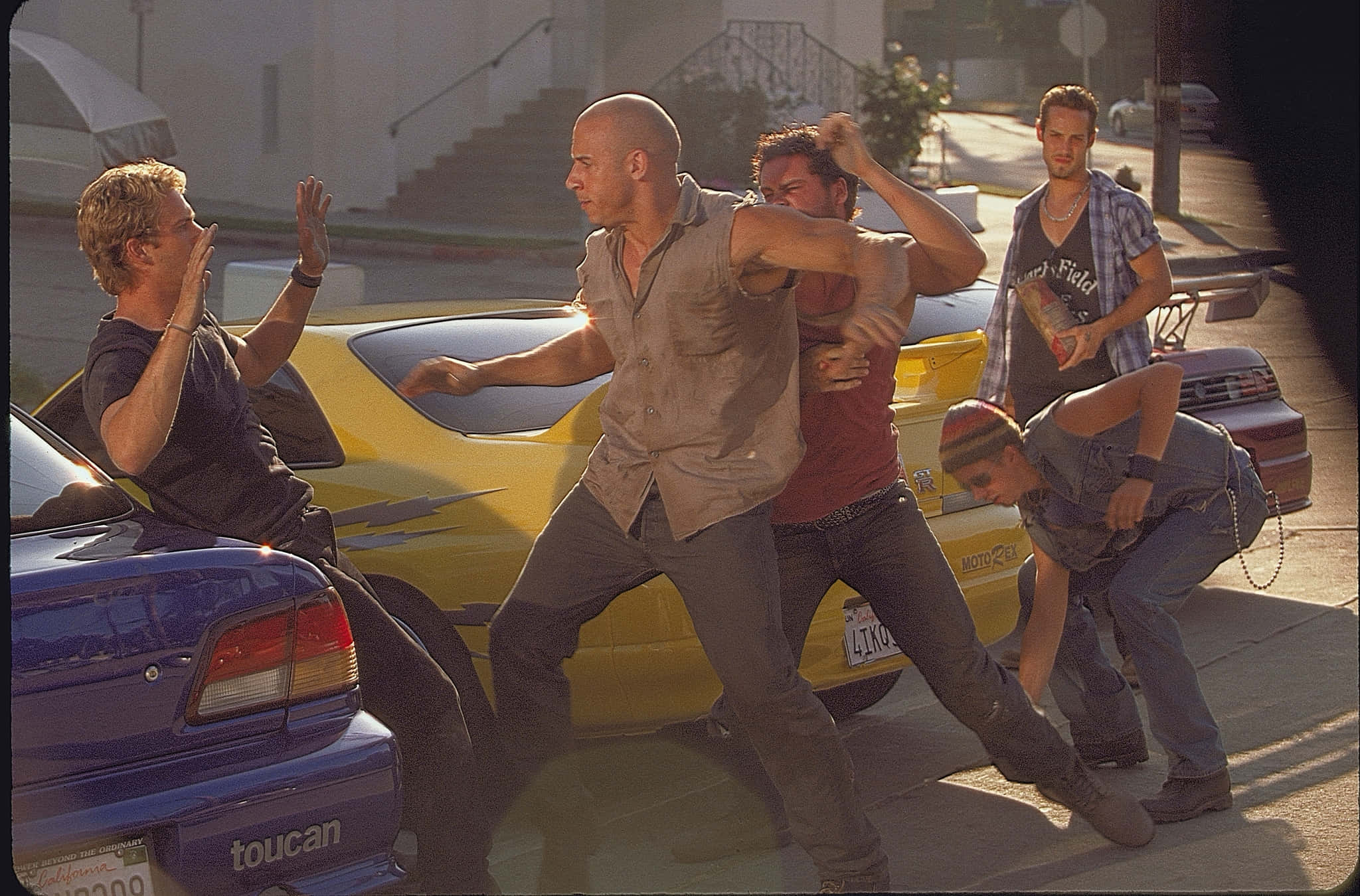 Fast And Furious 1 桌布 壁紙 — 一群男人在陽光明媚的街道上，在停泊的汽車旁邊打鬥，其中一名男子坐在黃色汽車內，處於混亂的中心。
