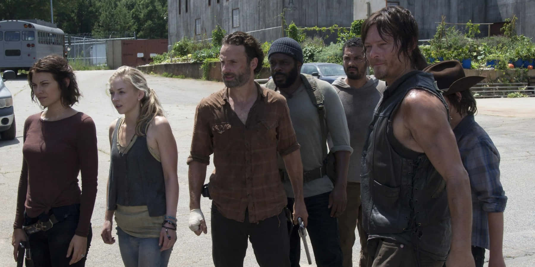 walking dead 桌布 壁紙 — 一群疲憊的倖存者走過一個陽光普照、雜草叢生的城市空地，背景有廢棄車輛和破舊建築物。