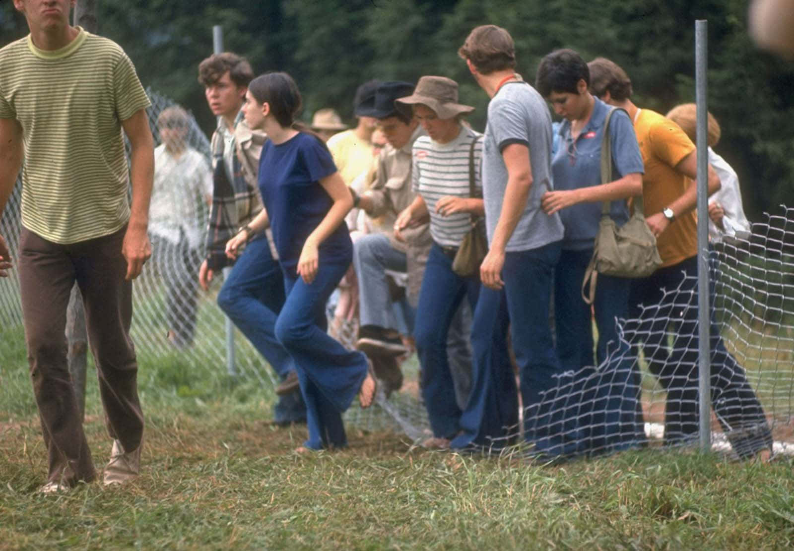 woodstock 桌布 壁紙 — 一群穿著1970年代服裝的年輕人沿著草地旁邊的破損鐵絲網圍欄行走。